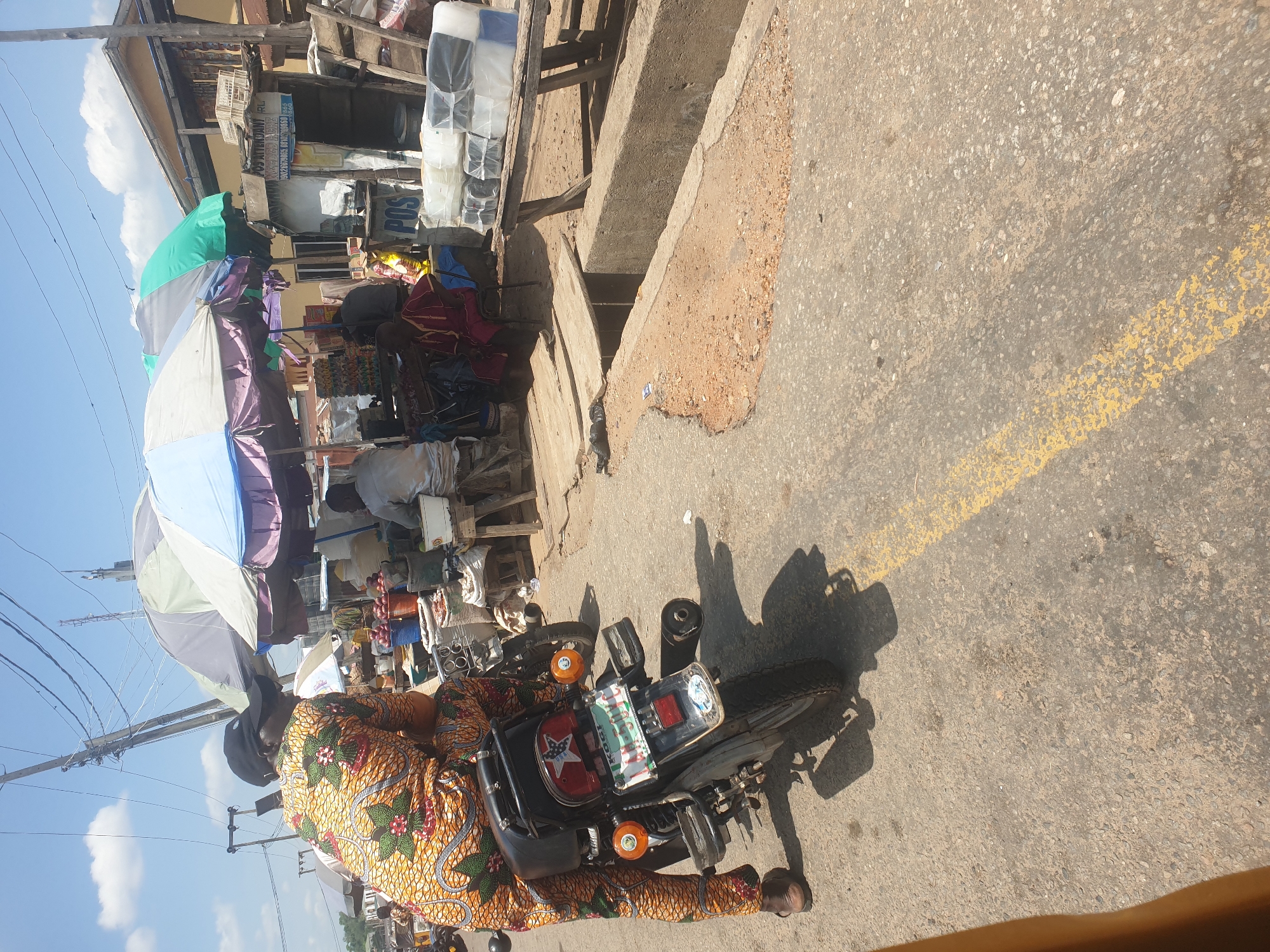 The image depicts a street scene in Lokoja, Nigeria. A person wearing a colorful patterned fabric is standing near a parked motorcycle on a paved road. The road has a faded yellow painted line running along it. The motorcycle has a license plate and an additional red star-shaped reflector or emblem. Behind the motorcycle and person, there is a bustling street market with numerous vendors, umbrellas, and various goods on display. A building with signage is visible in the background. The overall environment suggests a busy commercial area.