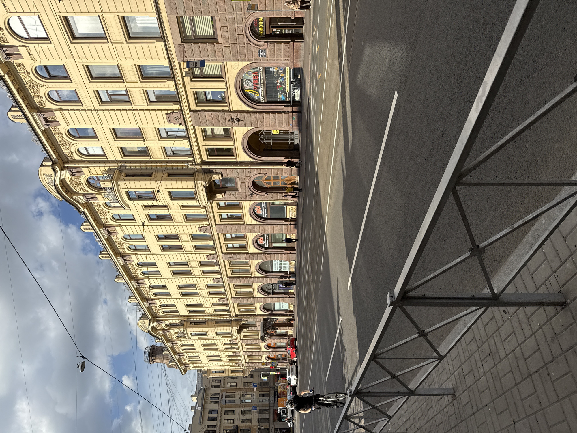 The image depicts a wide, paved street in Saint Petersburg, Russia, with a light-colored, ornate building lining the right side. The building features multiple stories with arched storefronts on the ground level and numerous windows on the upper floors, adorned with decorative elements. The street is marked with white lane lines and is relatively empty, with a few cars visible in the distance. A cyclist wearing dark clothing and a helmet rides a bicycle on the left side of the street, near a gray metal fence and a sidewalk paved with rectangular blocks. The sky is partially cloudy, casting shadows on the street and the building. Several overhead wires and a lamppost are visible against the sky. The overall scene suggests a typical urban environment in Saint Petersburg on a day with mixed weather conditions.