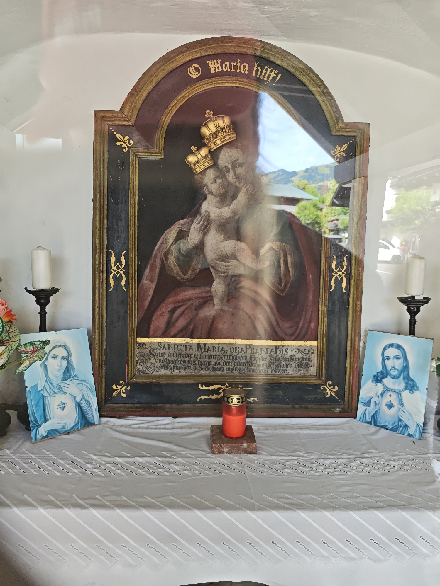 The image shows an indoor shrine or niche, likely located in Kitzbühel, Austria, given the context provided. The central focus is a framed painting of the Madonna and Child, both wearing crowns. Above the painting, in gold lettering on a dark background, is the inscription "Maria hilf!". Below the painting, there is a gold-colored decorative border framing a text that is partially obscured by glare and reflection but appears to be in Latin.
To the left of the central painting, a framed image of the Virgin Mary, depicted in blue and white, is propped up. To the right, a similar framed image of Jesus Christ is also propped up, with his hands held over his chest.
In front of the central painting, a red votive candle in a glass holder sits on a small marble slab. Two white pillar candles in dark, ornate candle holders are positioned on either side of the shrine, flanking the framed images. The shrine is covered by a white tablecloth with a decorative, patterned overlay. The background wall is white, and the overall lighting suggests an indoor environment with some natural light coming in, causing reflections on the glass of the central painting. The presence of religious imagery and votive candles indicates this is a devotional space.