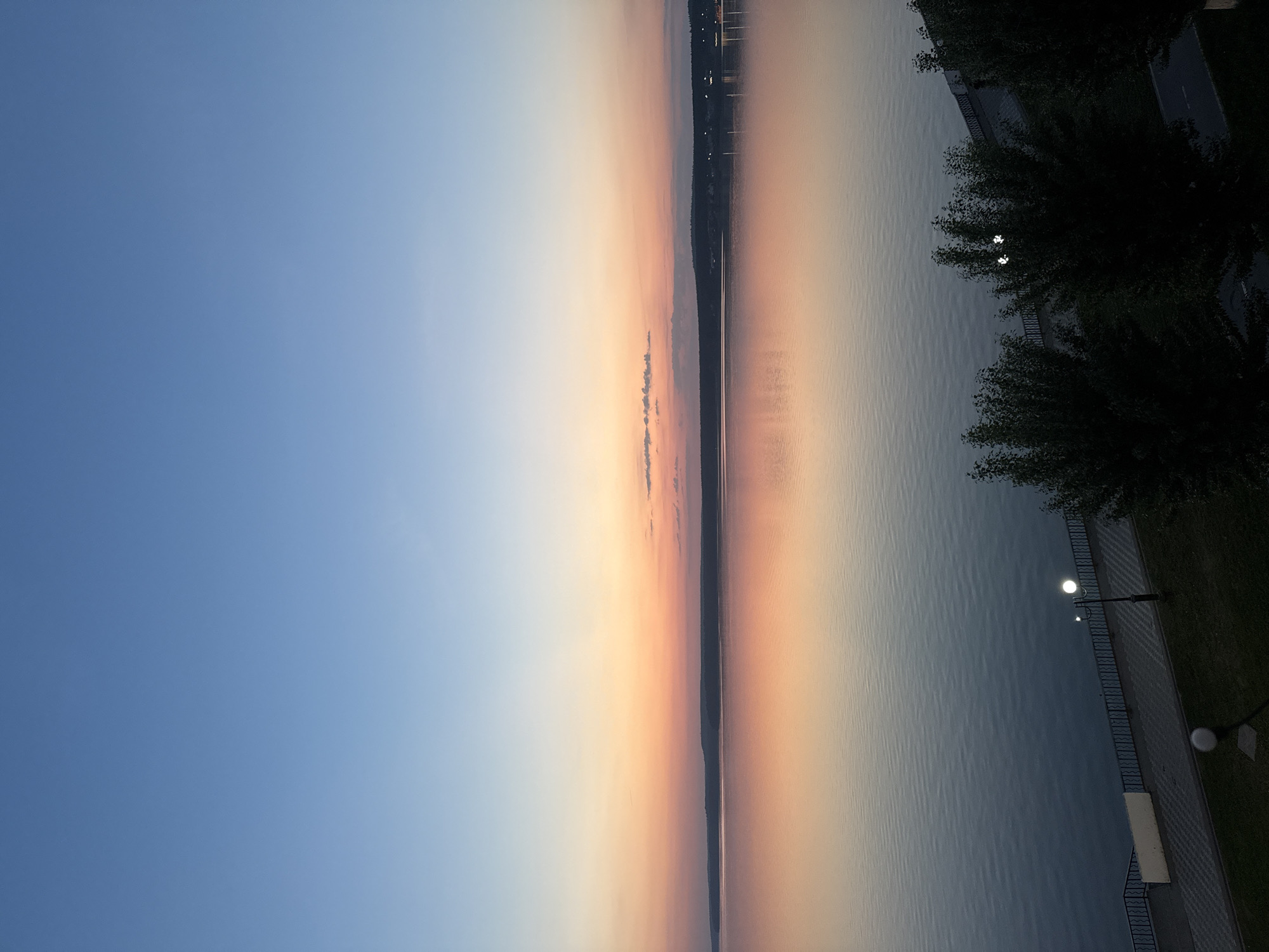 The image depicts a landscape at sunset or sunrise in Votkinsk, Russia. The sky is a gradient of blue to orange. The long body of water is likely the Votkinsk Reservoir, with its surface reflecting the colors of the sky. Along the shoreline are trees and a paved area with street lights. The precise location within Votkinsk would be along the bank of the reservoir.