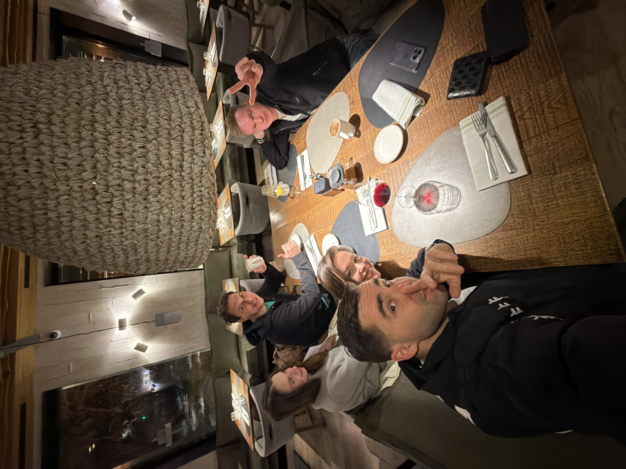 The image, rotated 90 degrees clockwise, depicts six individuals (four males, two females) dining or socializing around a long wooden table in what appears to be a restaurant or upscale bar. The setting is indoors, with dim ambient lighting suggesting an evening or nighttime gathering.
The group is seated along one side of the table, which is set with individual grey oval placemats, white striped napkins, and cutlery. Various drinks are visible, including glasses with red wine and cocktails with yellow garnishes, as well as coffee cups. Personal items such as a black smartphone and a black wallet are also on the table.
On the left side of the frame, a man holds a cup and smiles. Next to him, a man in a dark hoodie looks towards the camera with fingers near his mouth. A woman with long brown hair smiles beside him. Further down, another woman with dark hair is partially visible. Across the table, a man in a black jacket is making a "peace" or "V" sign towards the camera, and another man leans on the table, looking forward.
The background features modern interior design with light-colored walls, recessed lighting fixtures, and a large window reflecting outdoor lights and structures. A large, textured, woven decorative element or light fixture occupies the left foreground. The overall atmosphere appears relaxed and convivial. No discernible text is visible within the image.