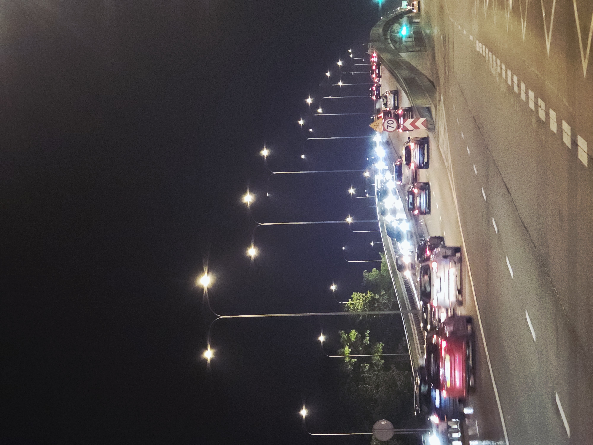 The image captures a nighttime scene of a highway in Warsaw, Poland, specifically showing traffic on a bridge. Cars are lined up, mostly in the right lane, with their headlights and taillights illuminated. Streetlights are visible along the highway, casting a glow on the road. A sign indicating a speed limit of 70 kilometers per hour is present. The sky is dark, and some greenery can be seen alongside the road, suggesting the presence of trees or other vegetation. The road markings and lane divisions are clearly visible.