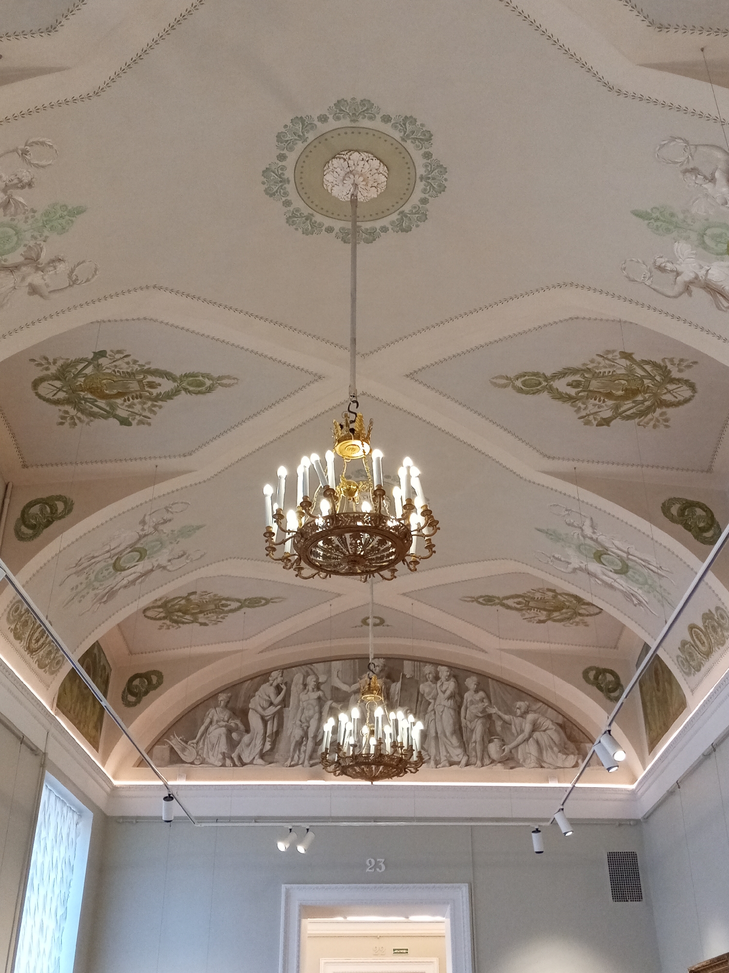 The image shows the interior of a building, specifically a room with an ornate ceiling. The ceiling is decorated with painted designs, including floral motifs, wreaths, and classical figures. Two chandeliers hang from the ceiling, each with multiple lights. The lower portion of the image includes a wall with a painted frieze depicting classical figures, lighting fixtures, and a doorway marked with the number "23." The style of the architecture and decoration suggests a location in Saint Petersburg, Russia, known for its classical and baroque buildings.
