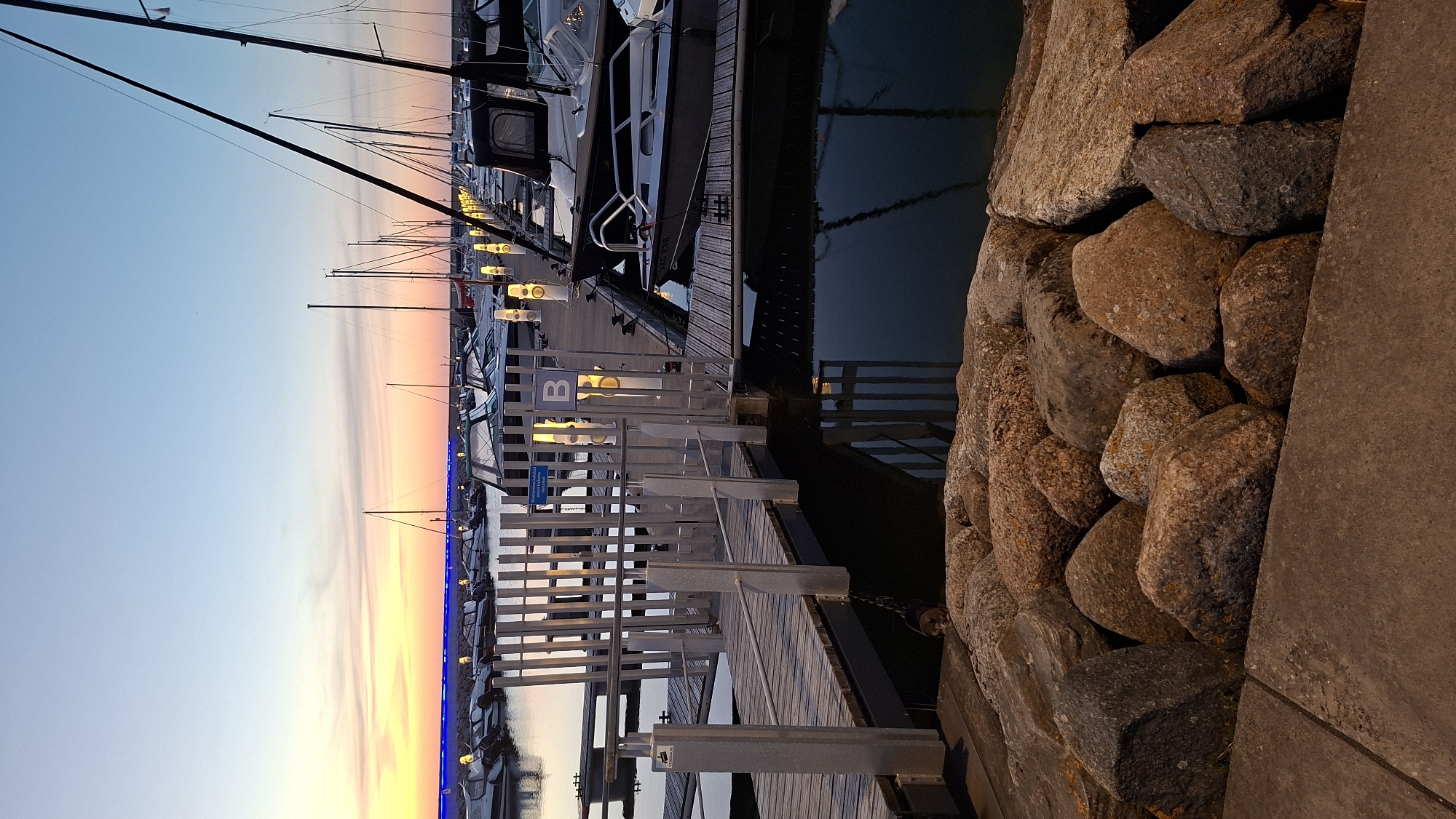The image depicts a marina at dusk, with several boats docked along a pier. A wooden walkway with metal railings leads to the pier, where a sign with the letter "B" is visible. The sky exhibits a gradient of colors, transitioning from a soft orange and pink near the horizon to a clear blue overhead. The water reflects the sky and the lights of the marina. In the foreground, large rocks and a concrete path are visible. The overall atmosphere is serene and tranquil. The scene is of the Pirita Marina in Tallinn, Estonia, a popular location for boating and water activities.
