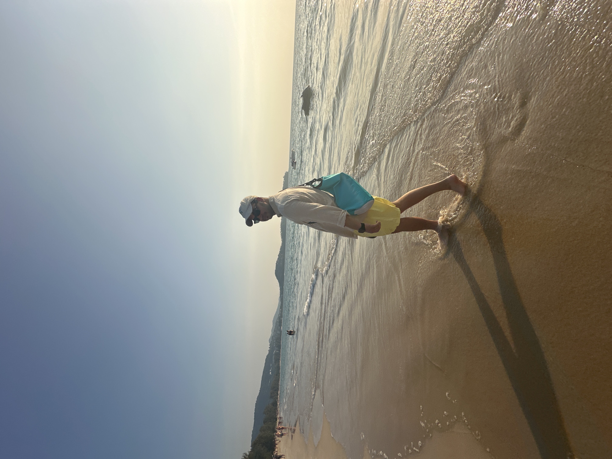 A male subject is captured walking barefoot along the wet sand of a beach in Choeng Thale, Thailand, during late afternoon or early morning. He wears a light-colored long-sleeve shirt, yellow shorts, a white baseball cap, sunglasses, and a black watch on his left wrist. A light blue dry bag is slung over his right shoulder. The subject is partially turned to his right but looks back towards the camera over his left shoulder. Small waves lap around his feet, creating foam on the shoreline. To the left, numerous individuals are visible walking along the extensive sandy beach, backed by lush green hills or mountains. To the right, the ocean stretches to the horizon, where the sky exhibits a warm, golden hue. Distant watercraft, including a jet ski, are discernible on the water. Long shadows extend from the subject across the sand towards the left side of the frame, indicative of a low sun angle.