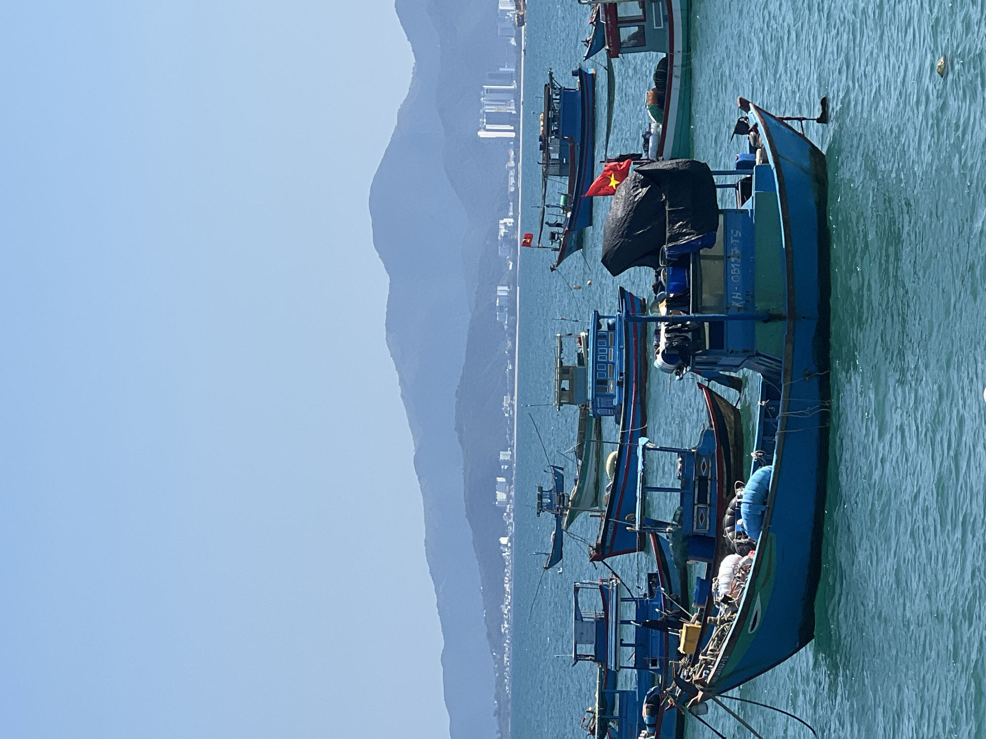 The image displays a wide shot of a harbor in Nha Trang, Vietnam, under a clear, light blue sky. Numerous traditional wooden fishing boats, predominantly blue and green, are anchored on calm, turquoise water. The boat closest to the viewer, positioned slightly right of center, is blue with green trim and features the registration "KH-0817-TS" on its side. A black tarp covers a portion of this boat, and a red flag with a yellow star, identifiable as the national flag of Vietnam, is affixed to its stern. Other similar fishing vessels are scattered throughout the midground, some also displaying Vietnamese flags. In the background, a distinct range of mountains extends across the horizon, with a developed cityscape, including several high-rise buildings, visible along the distant coastline at the base of the mountains. No individuals are depicted in the scene.