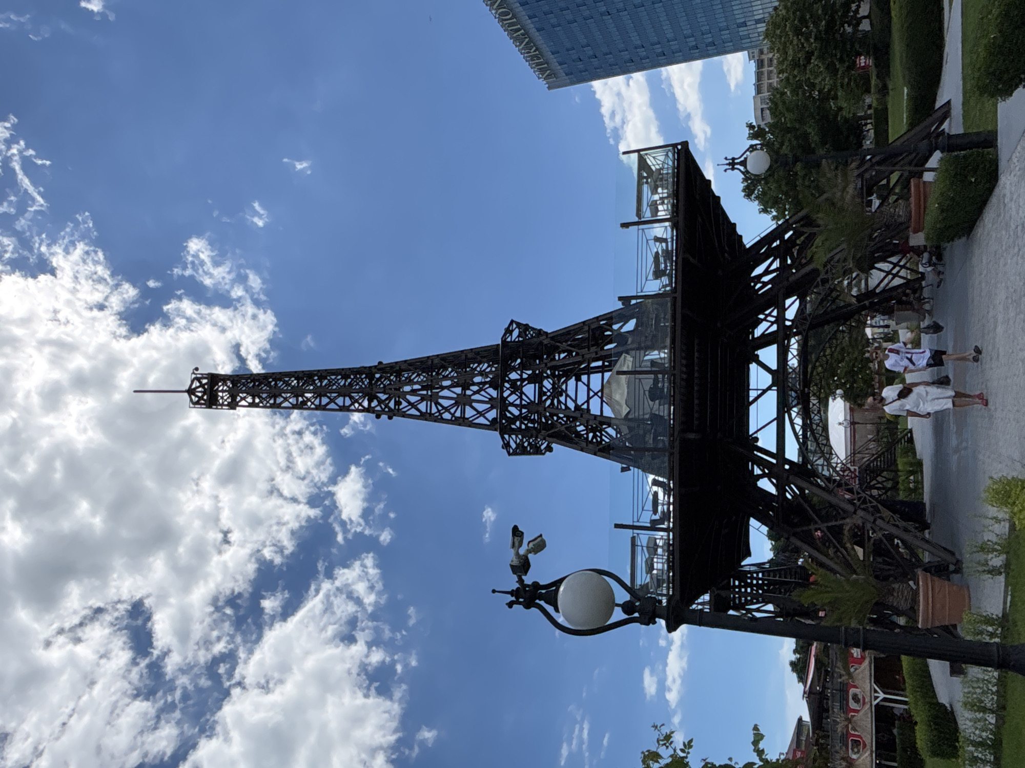 A replica of the Eiffel Tower, made of dark metal, stands prominently in an outdoor urban setting. The replica features a viewing platform with glass railings, furnished with tables and chairs, suggesting it may be a restaurant or bar area. A traditional street lamp with a globe light is attached to the structure. In the background, a modern glass-fronted skyscraper with the word "INTERNATIONAL" visible on its facade, signifies an urban environment. The sky is a clear blue with scattered white clouds. In the foreground, a paved walkway runs in front of the tower, with neatly trimmed green bushes and potted palm trees lining the path. Two individuals, a woman in a white dress and a man in a white t-shirt and shorts, are walking away from the camera on the path. The location is identified as Varna, Bulgaria.