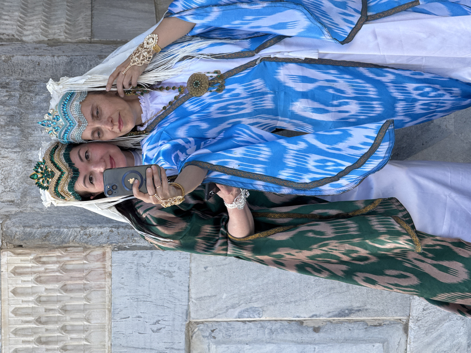 Two women are in Samarkand City, Uzbekistan, posing for a selfie. The woman at the bottom of the frame holds a smartphone, and wears a green and beige ikat dress with a gold trim, and a traditional headdress with green and gold jewels. The woman above wears a white dress with a blue and white ikat robe, a jeweled headdress with blue stones, and a jeweled bracelet. They are in front of a stone structure with decorative molding.