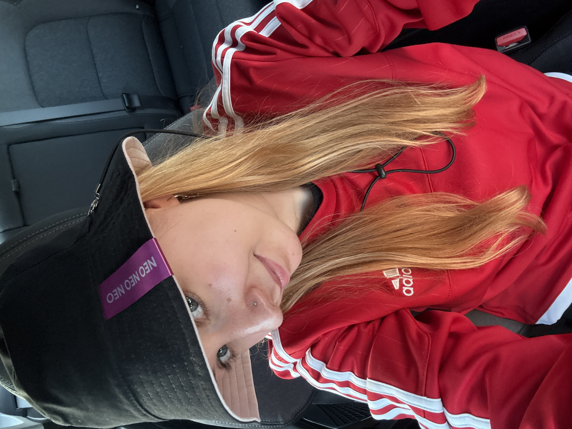 The image depicts a fair-skinned individual with blonde hair, wearing a black bucket hat with a purple tag that says "NEO NEO NEO" and a red Adidas tracksuit. The individual has a nose piercing and is facing the camera. The person appears to be sitting in a car, as indicated by the seat and seatbelt visible in the background.
Without further information, it is impossible to determine if the image was taken in Minsk, Belarus.