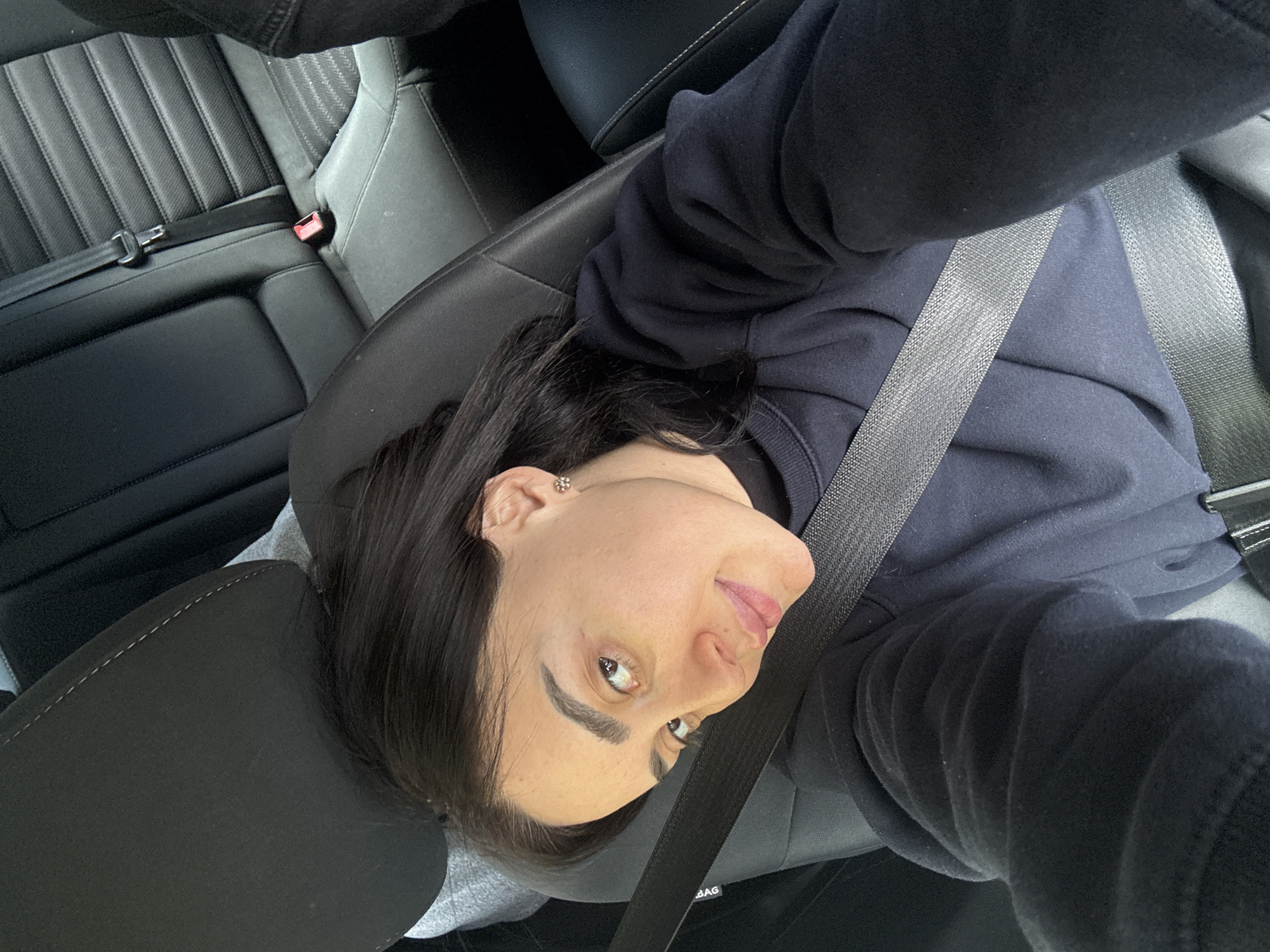 A person wearing a dark sweatshirt and buckled into a seatbelt is in the backseat of a car. The seat is black leather, with a gray headrest. The back seats are vertically ribbed. The setting is unknown, so it's impossible to determine the location in Tbilisi, Georgia.