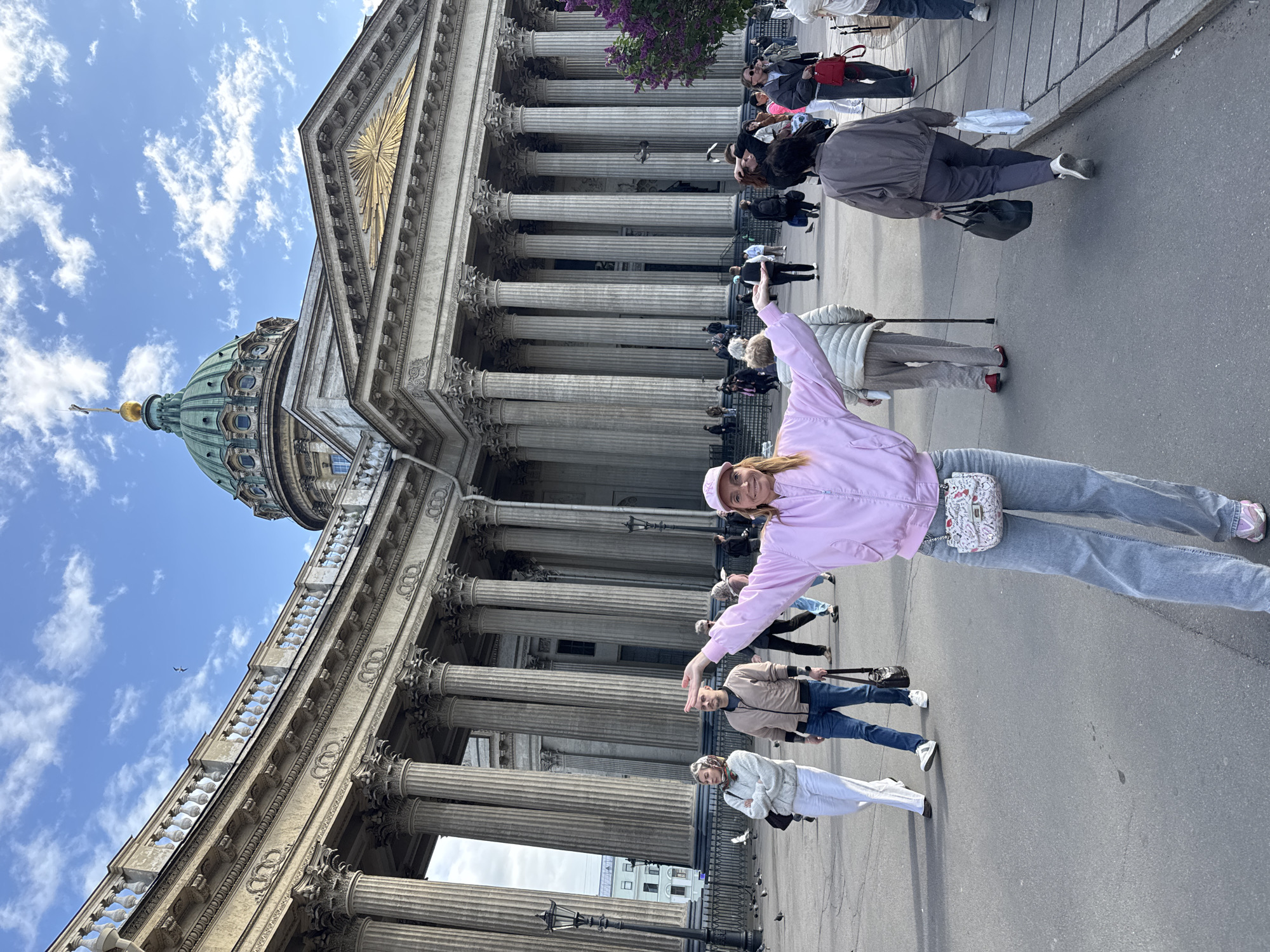 A medium shot depicts a pink-jacketed woman posing in front of the Kazan Cathedral in Saint Petersburg, Russia. The cathedral's facade is characterized by tall, gray columns supporting the structure, with a partial view of its green dome and a blue sky dotted with clouds above. The woman, positioned at an angle, has her arms outstretched, and appears to be smiling at the camera. Several other individuals are visible in the background, walking or standing near the cathedral. The ground appears to be paved with gray asphalt.