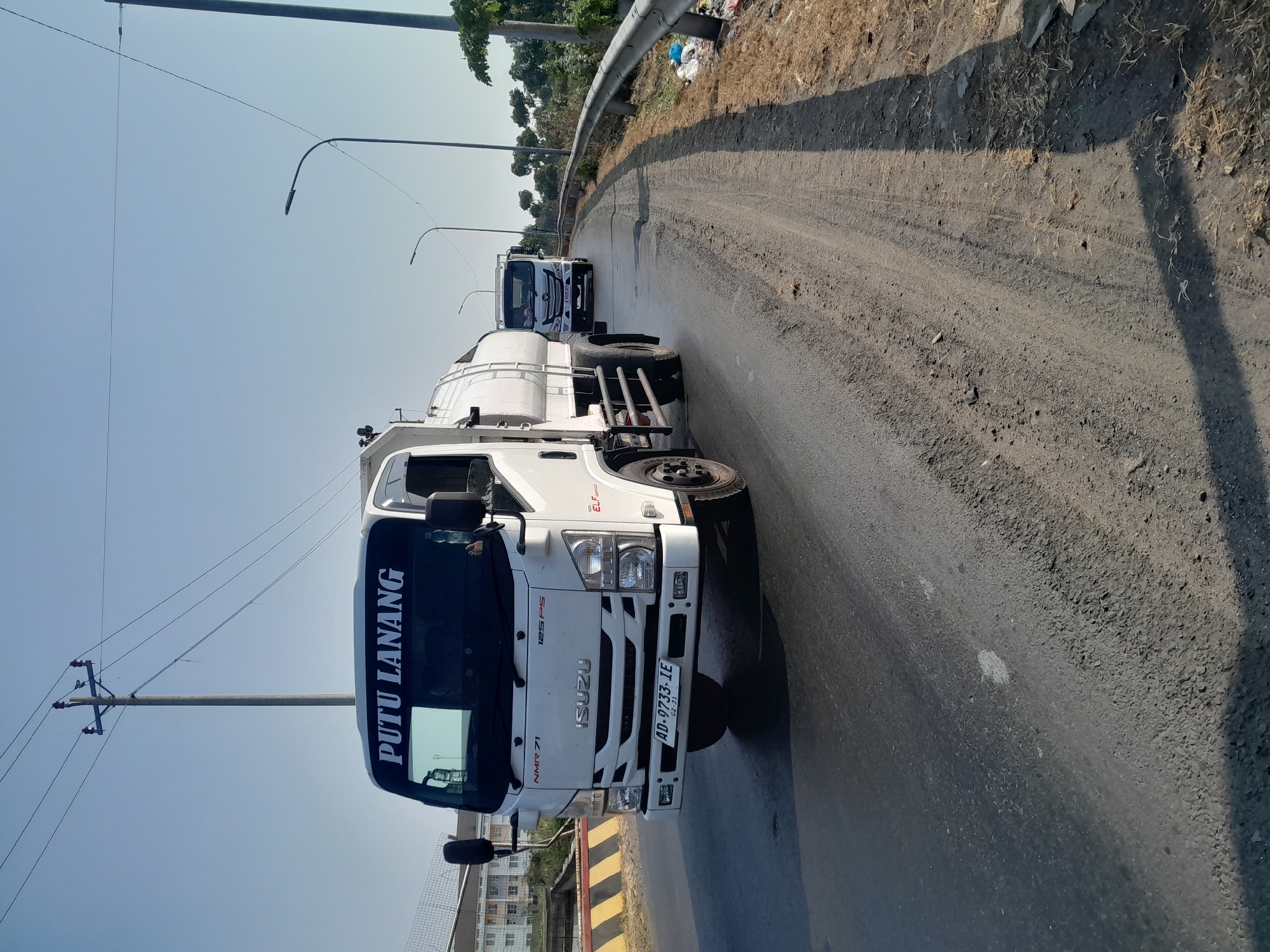 The image captures a sunny day on a road in Tegal, Indonesia. In the foreground, a white Isuzu NMR 71 truck with the text "PUTU LANANG" on its windshield is driving on the right side of the road. The truck has a white water tank attached to its rear. Its license plate reads "AD-9733-IE". The road surface is asphalt with some patches of gravel on the shoulder. In the background, another white truck is visible further down the road. A metal guardrail runs along the right side of the road, and beyond that, dry grass and trees can be seen. Power lines and utility poles are present overhead. A multi-story building is visible on the left side of the road. The sky is clear and blue.