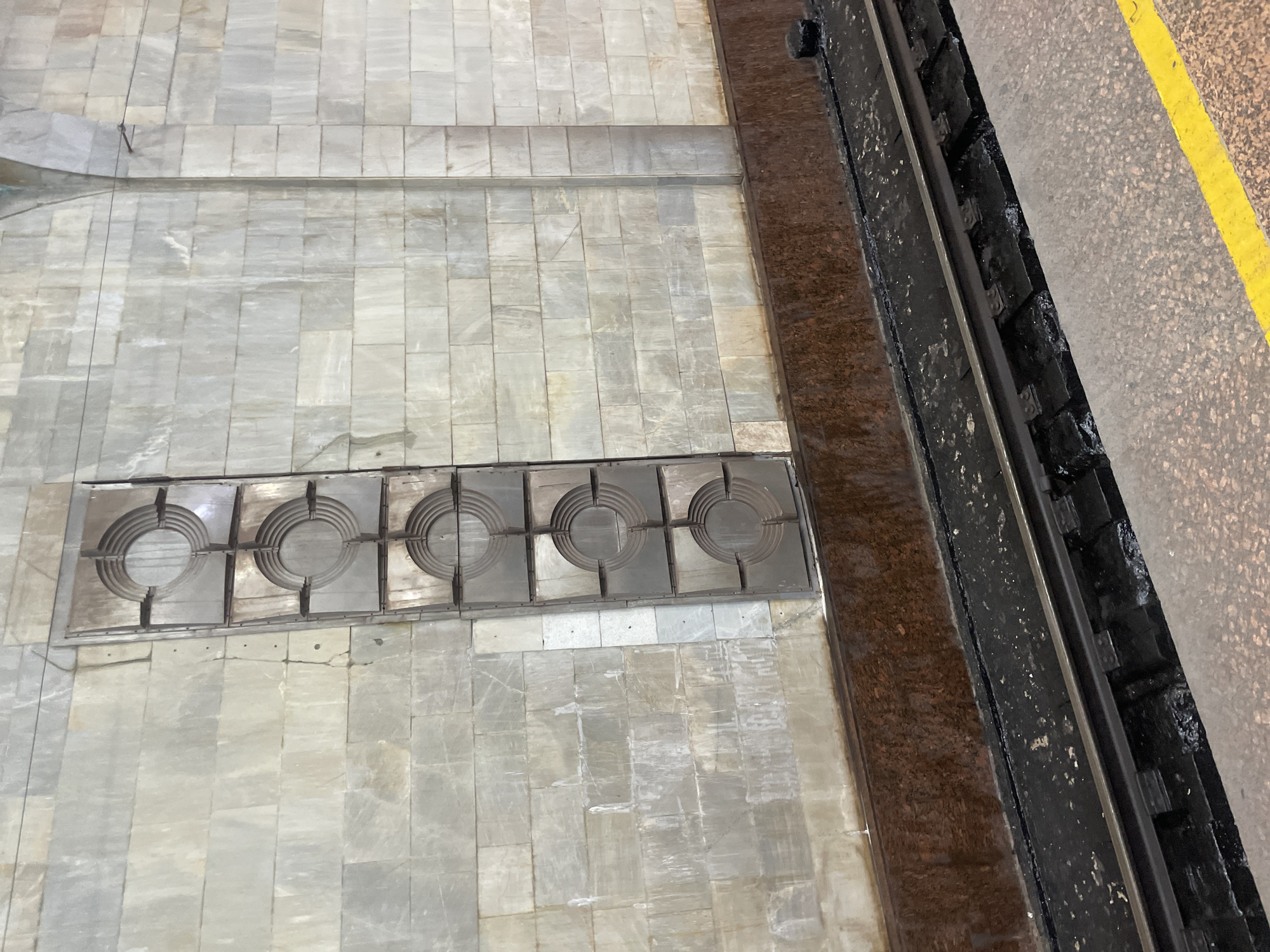 The image shows a wall in a Tashkent metro station, featuring a decorative panel composed of several circular segments. The wall itself is clad in light-colored marble tiles with visible signs of wear and staining. Below the wall are train tracks, with a dark, possibly asphalt or tar, base. A platform with a yellow line marking is visible in the foreground. The overall impression is of an older, somewhat weathered, urban transit environment.
