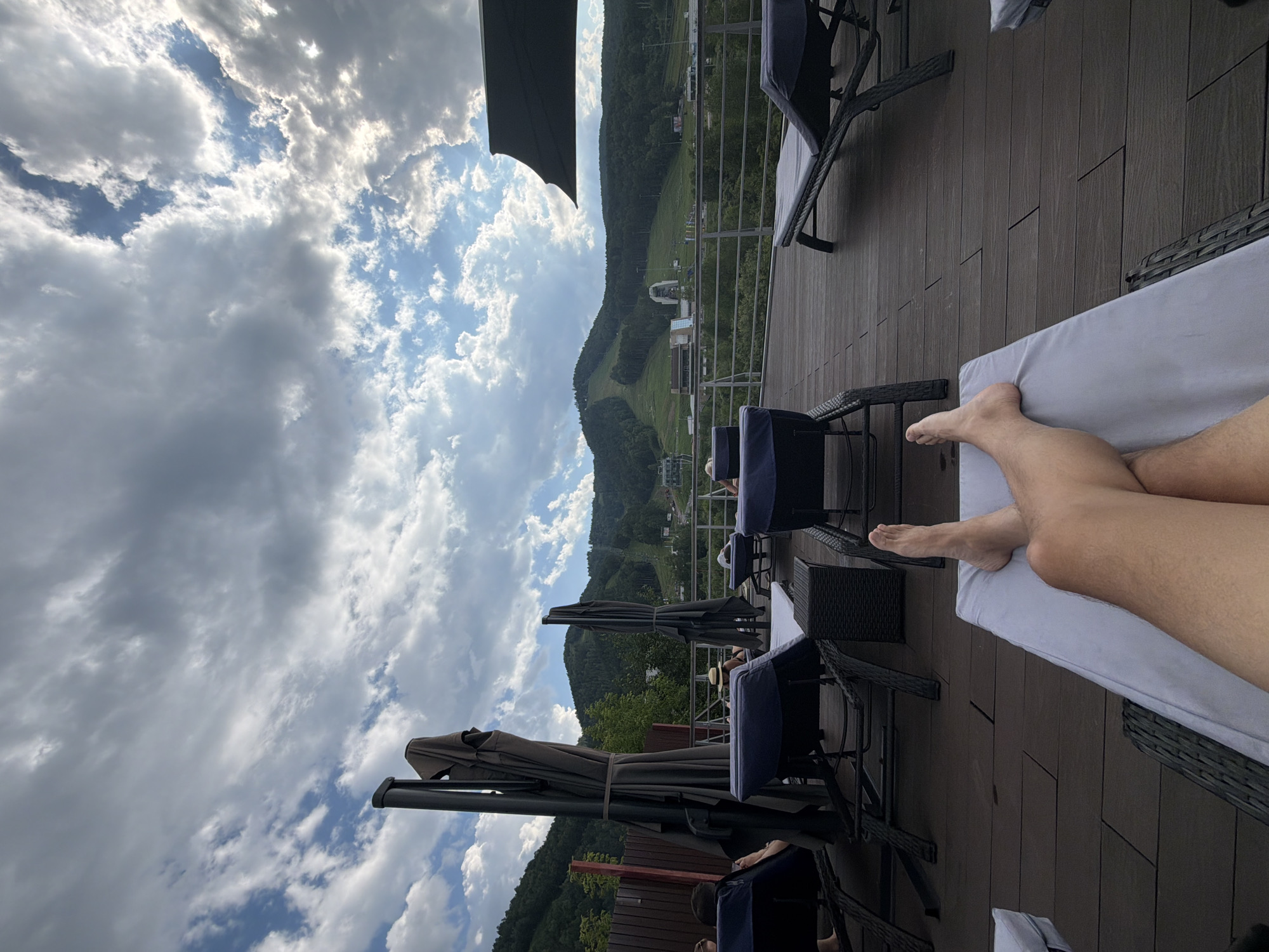The image shows a first-person perspective of a person relaxing on a lounge chair on a wooden deck. The person's legs and feet are visible, crossed at the ankles, and they are lying on a white cushion. In the background, there are several other lounge chairs, some occupied by people, and large dark gray umbrellas, some folded. Beyond the deck, there is a railing overlooking a grassy hillside with a ski resort area. The ski resort features buildings, ski lifts, and a sloped area with tracks. The hillside is covered with trees, and further in the distance, more mountains are visible under a cloudy sky with patches of blue. The overall scene suggests an outdoor relaxation area with a scenic view of a mountain landscape and ski facilities. The location is not Krasnoyarsk, Russia, as it depicts a ski resort in a mountainous region, which is not characteristic of Krasnoyarsk.