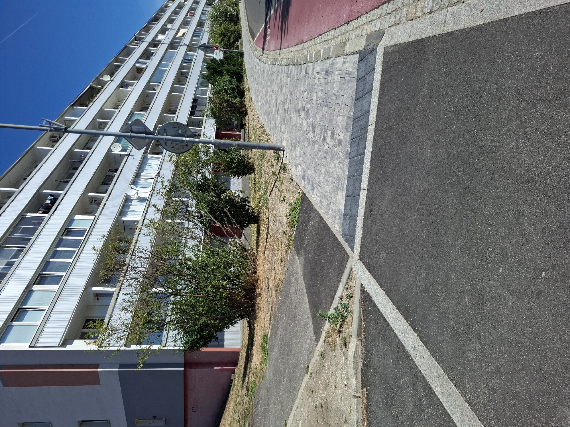 This is a photograph of an apartment building in Bratislava, Slovakia, taken on a sunny day. The building appears to be multi-story with a light-colored facade and numerous balconies. In the foreground, there is a paved walkway with distinct sections of interlocking pavers and asphalt. A red-colored lane, possibly for cycling or other traffic, runs alongside the walkway, separated by a granite curb. Green bushes and trees are visible between the building and the walkway. A metal pole with traffic signs is also present. The ground in front of the building is covered with asphalt and gravel, with some patches of dry grass. The sky is clear and blue.