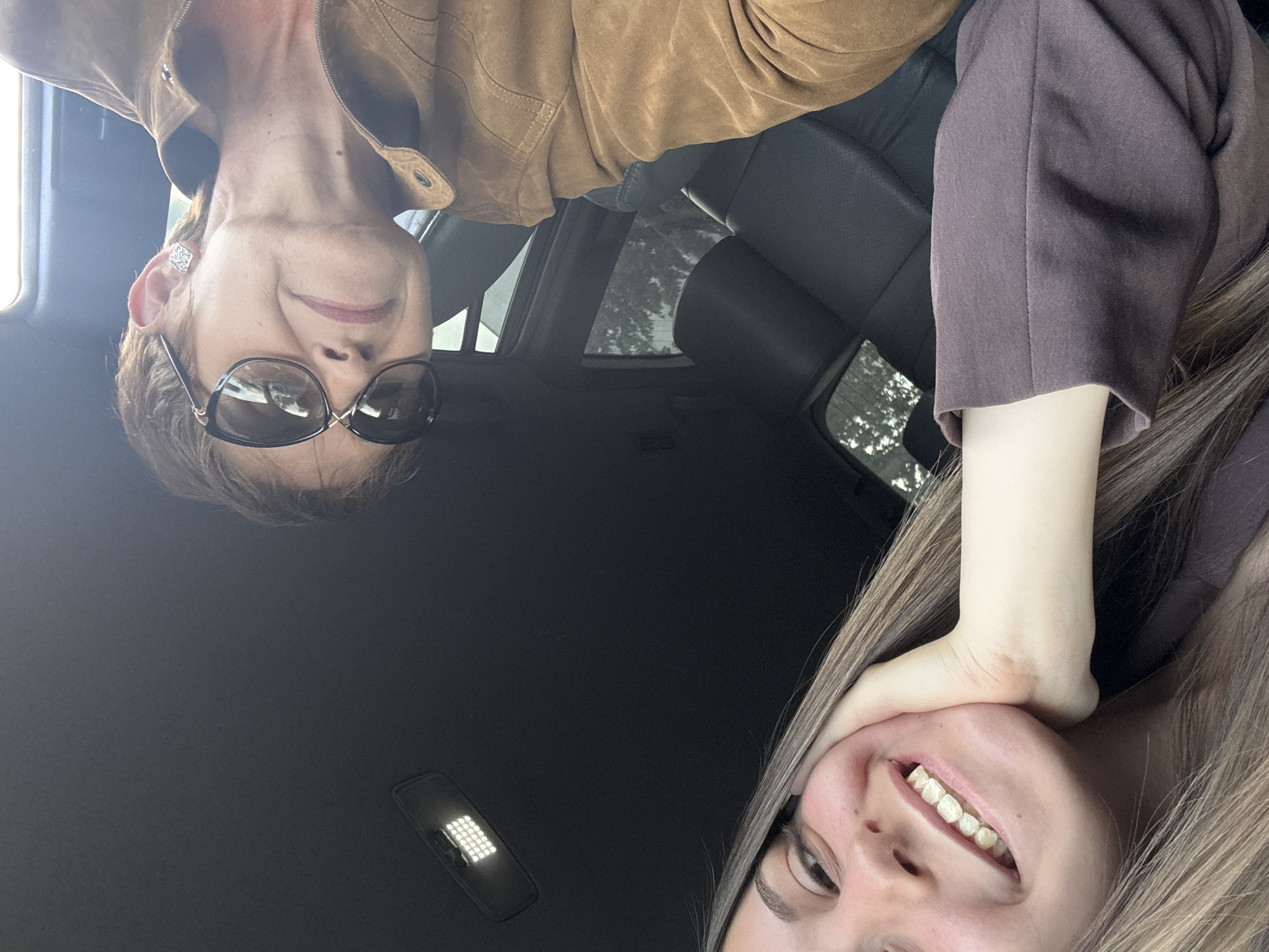 The image appears to be a selfie taken inside a car, possibly a photograph from the roof of the interior looking down. Two individuals are visible: an older woman wearing a brown suede jacket and sunglasses, and a younger woman in a brown shirt or jacket, touching her face. The car's interior, including seats, a window showing greenery outside, and a dome light, can be discerned. It is not possible to determine if the location is Burgas, Bulgaria, with the information given.