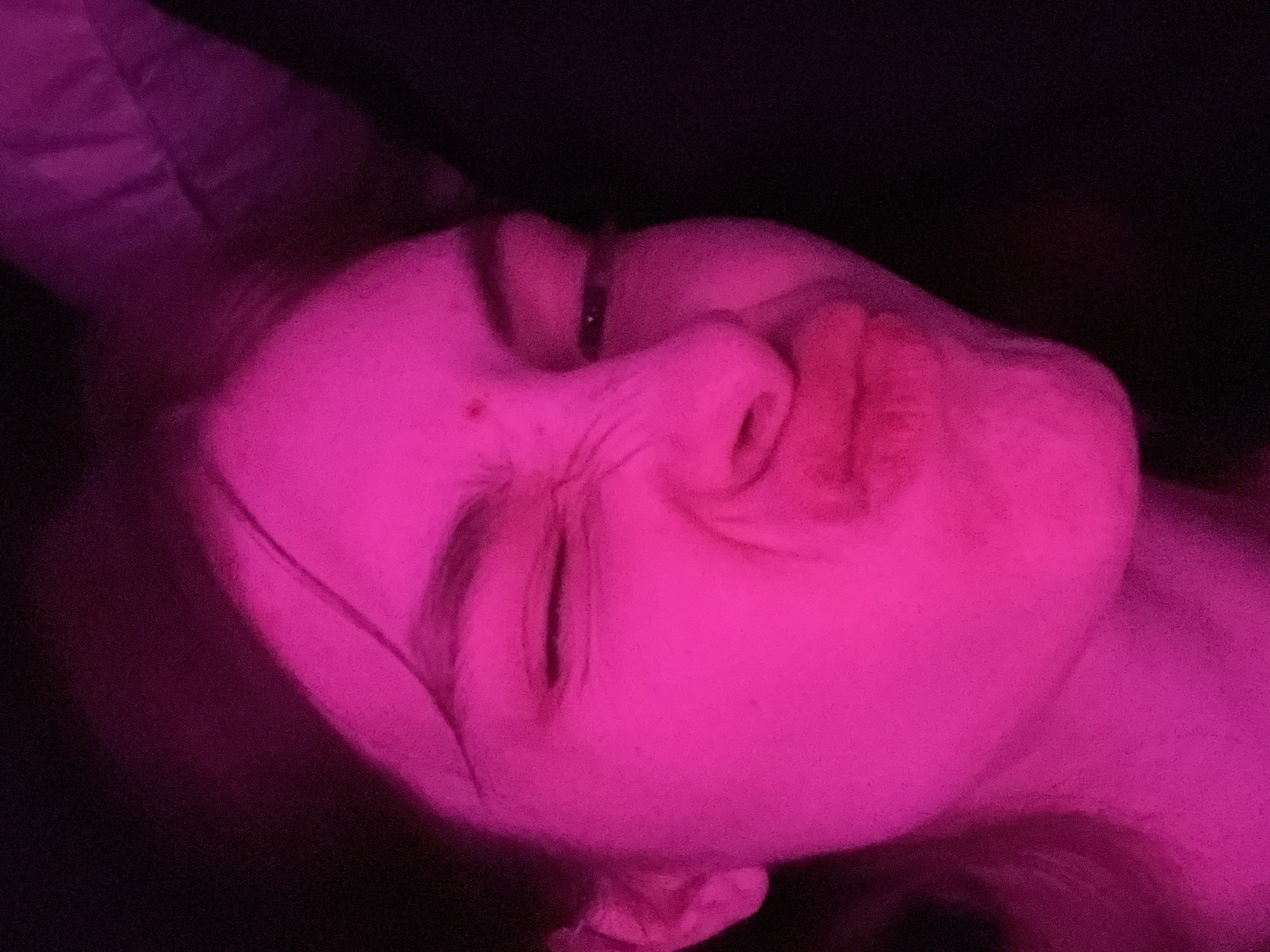 A close-up, upward-angled view shows a person's face illuminated by a pink light. The individual is lying down, with their head resting on what appears to be a pillow or soft surface. Their eyes are mostly closed, and their expression appears relaxed. The background is dark and undefined. There is no indication of the location being Saint Petersburg, Russia.