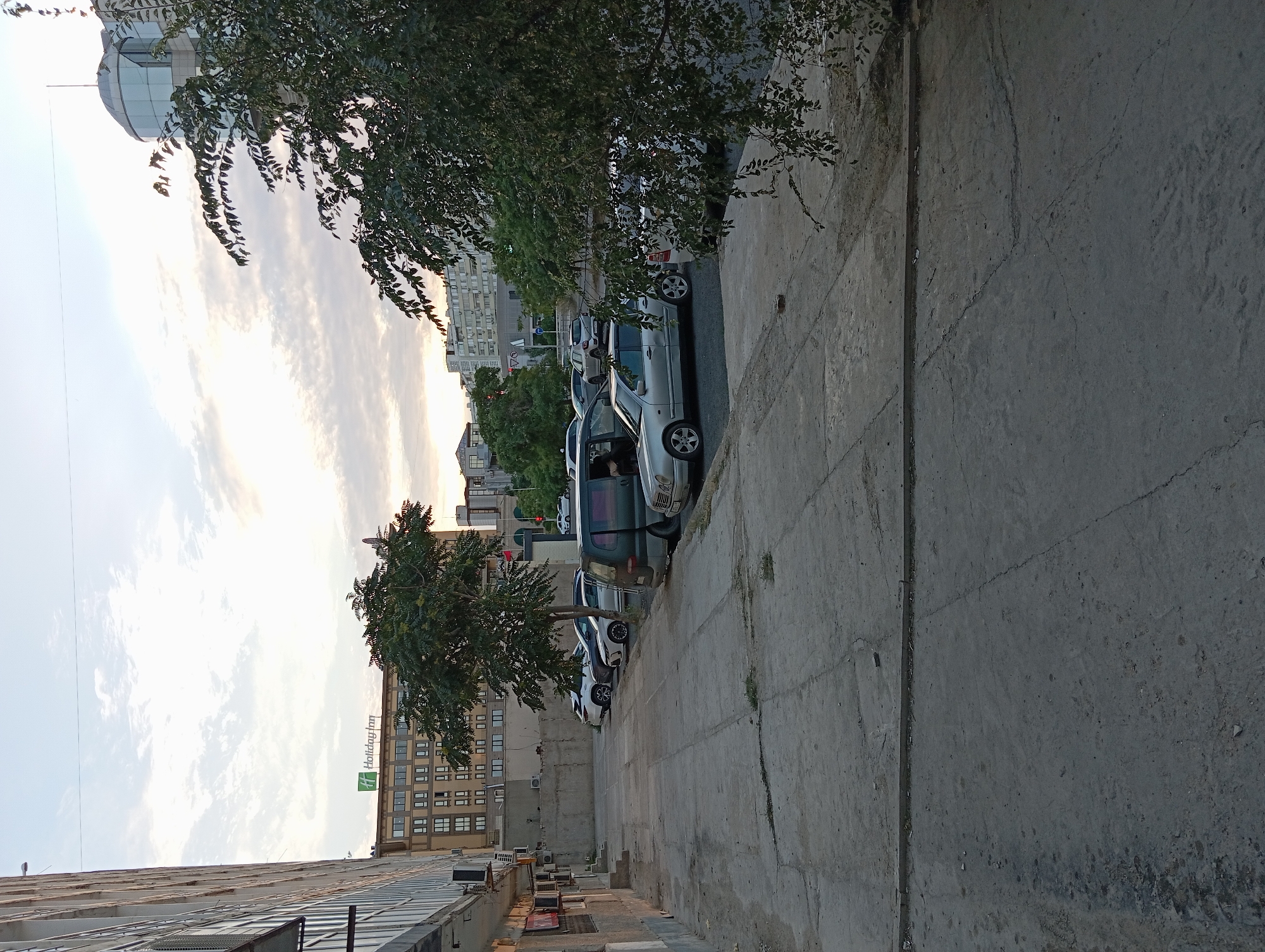 The image depicts a parking lot in front of a building with "Holiday Inn" signage, indicating a commercial area likely within the city of Aktau, Kazakhstan. Several cars are parked in the lot, including a silver sedan, a white SUV, and a dark-colored van. The buildings in the background appear to be a mix of modern and older structures, with trees interspersed throughout the urban landscape. The sky is cloudy, suggesting it may be late afternoon or early evening. The foreground shows a concrete pavement with some debris, typical of an outdoor urban setting.