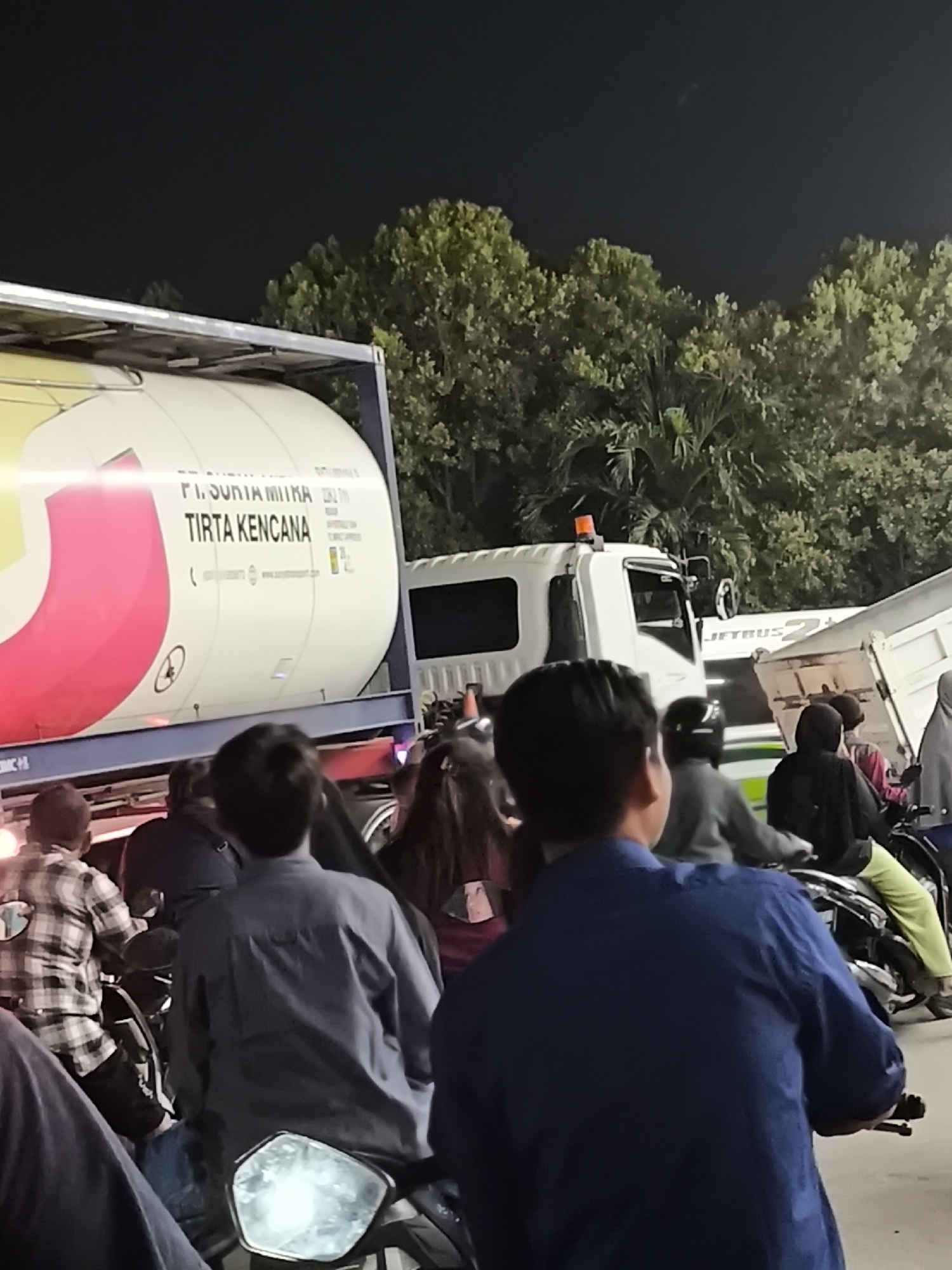 In the city of Waringinkurung, Indonesia, at night, a large white tanker truck with the text "PT. SURYA MITRA TIRTA KENCANA" is visible. The truck is parked, and several people on motorcycles are gathered around it. The individuals are mostly seen from the back, wearing casual clothing, some with helmets. The environment is dimly lit, with trees visible in the background and the sky appearing dark. The scene suggests a possible roadside gathering or a temporary stop for the truck.