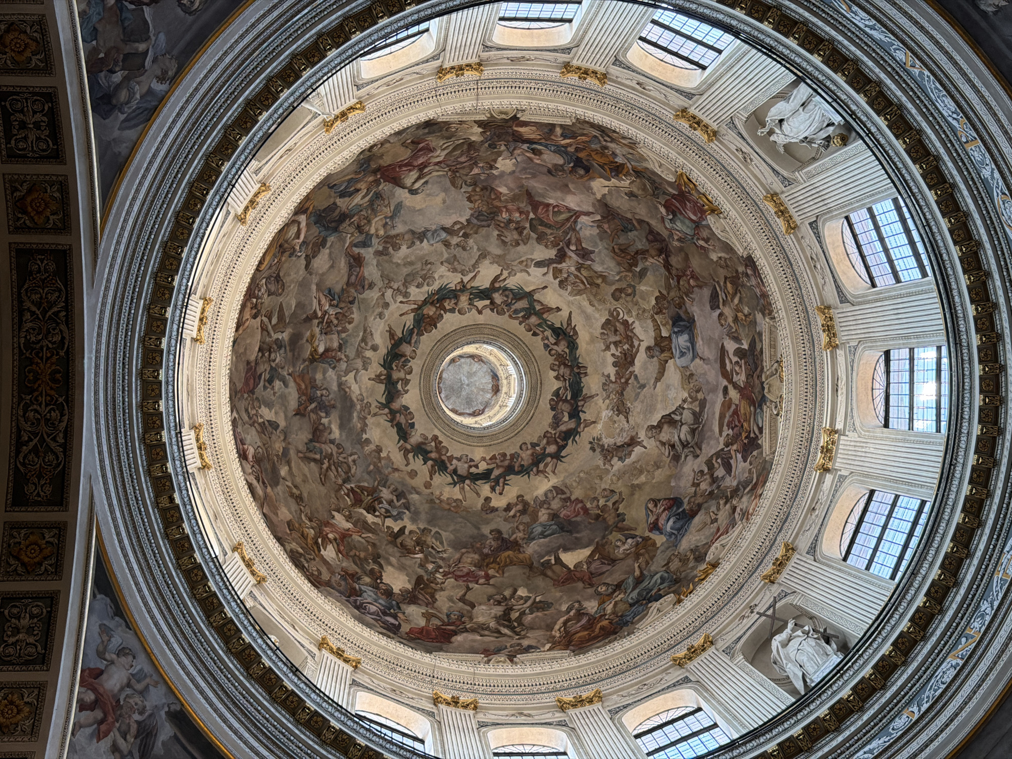 The image is an upward view into the dome of the Basilica of Sant'Andrea in Mantua, Italy. The dome's interior is richly decorated with a fresco depicting a multitude of figures, possibly religious or mythological in nature, swirling upwards toward a central oculus. Around the base of the dome, windows punctuate the architecture, providing natural light. Sculptural details, along with gilded decorations, accentuate the architectural features. The image is taken from a perspective looking directly up, emphasizing the dome's curvature and intricate artistry.