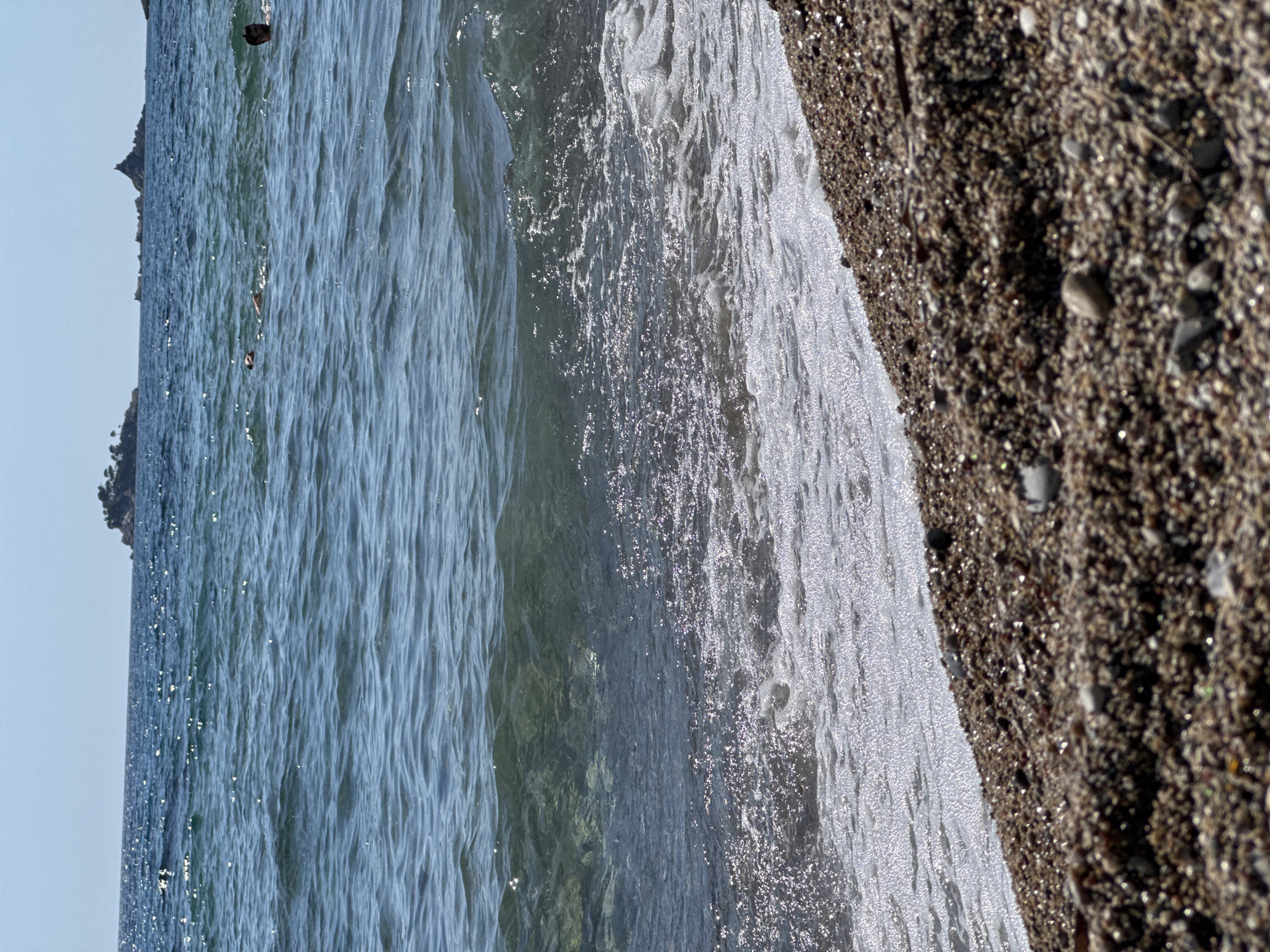This is a close-up, eye-level, vertical shot of a pebbly beach in Đurovići, Montenegro, with the sea and sky dominating the background. The foreground consists of the pebbly shore, with various shades of brown, gray, and white stones clearly visible, some appearing wet and glistening. The sea is depicted with waves gently lapping onto the shore, creating foamy white patterns. The water itself is a clear, deep blue, with sunlight reflecting off its surface, creating sparkling highlights. In the mid-ground, several people are visible swimming or floating in the water. Further in the background, there are several rocky islets or small islands, some with sparse vegetation, set against a clear, pale blue sky. The overall impression is a tranquil beach scene on a sunny day.