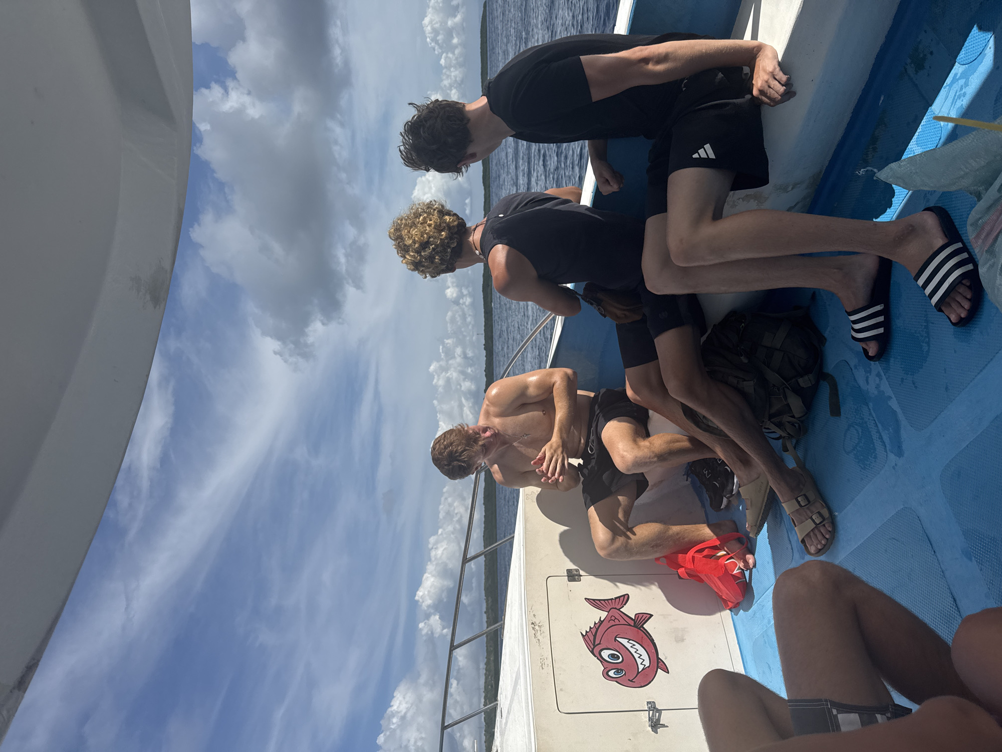 Several people are on a boat, looking over the edge into the water. Two people lean over the side of the boat towards the rear, followed by a bare-chested individual facing backwards toward the stern with his hands clasped in front of him. The environment consists of a bright, cloudy sky and dark water. The boat appears to be light blue inside and white outside. Nusa Penida, Indonesia, is located in the water.
