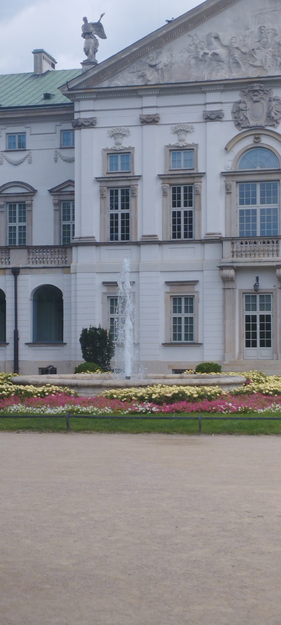 This is a medium-shot, eye-level, vertical-orientation photograph of the Royal Castle in Warsaw, Poland. The castle is a large, white, Baroque-style building with a green roof. In the foreground, there is a circular fountain with water spraying upwards. Around the fountain is a bed of colorful flowers, with pink, white, and yellow blossoms. A gravel path is visible in the lower portion of the image. The building has multiple windows, some of which are arched, and decorative sculptures above the upper floor. A statue of an angel is visible on the roofline. The sky is overcast.