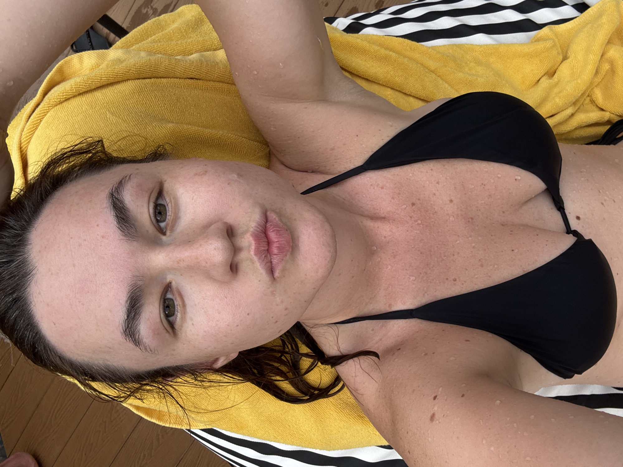 A woman is lying on a yellow towel on a wooden deck. She is wearing a black bikini top and her hair is wet. Her face is turned towards the camera with her lips pursed. Water droplets are visible on her skin and hair. The background shows a portion of the wooden deck and the striped edge of the towel. The image appears to be taken in Chalong, Thailand.