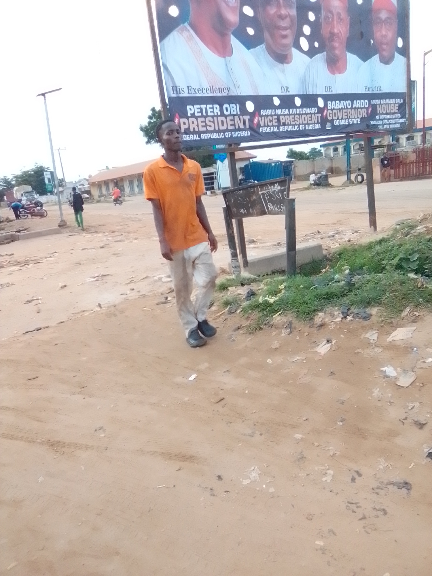 A young man in an orange t-shirt and light-colored pants walks on a dirt road in Gombe, Nigeria. He is facing towards the left of the frame and appears to be looking at something out of view. In the background, a large billboard displays images of political figures, including Peter Obi and Babayo Ardo, with text indicating their political positions such as President and Governor of Gombe State. The environment is dusty and appears to be an urban or semi-urban area, with some buildings and scattered debris visible. A few other people and vehicles are present in the distance.