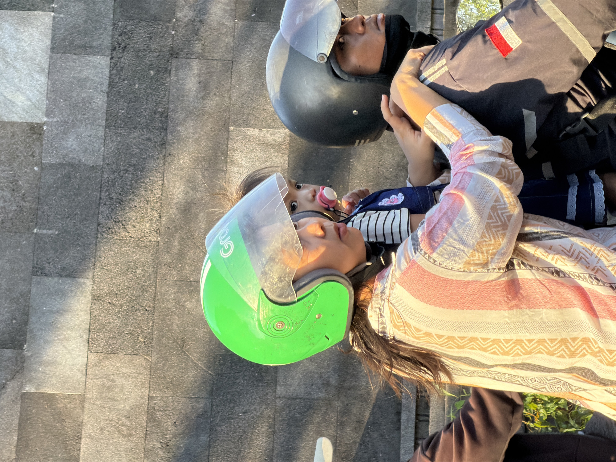 A woman, a child, and a man are depicted outdoors, likely on a motorcycle, viewed from the left side. The scene is illuminated by strong directional sunlight, casting shadows.
The woman is positioned centrally, wearing a bright green helmet with "Grab" branding visible on its right side. Her dark hair extends from beneath the helmet. She wears a striped shirt featuring vertical bands of white, pink, peach, and beige, with geometric patterns. Her face, partially obscured by the helmet's visor, is visible in profile, looking slightly downward and to the right. Her right hand is placed on the man's left arm.
Behind the woman and slightly to her right, a toddler-aged child is visible. The child has dark hair, looks towards the right, and has a pink pacifier in its mouth. The child wears a horizontally striped top and a blue garment, possibly overalls or a dress, beneath.
On the far right, a man, presumably the driver, wears a dark grey helmet with a clear visor. His face is seen in profile, looking straight ahead. A black neck gaiter covers his lower face. He wears a dark blue jacket or uniform with two horizontal white stripes on the sleeve and a rectangular patch on his upper left arm displaying the red and white horizontal stripes of the Indonesian flag.
The background consists of a wall constructed from rectangular, rough-textured, dark grey stone blocks. Green foliage is partially visible in the lower left corner. The presence of the Indonesian flag patch and the "Grab" helmet indicates the location is in Indonesia, specifically North Kuta as specified.