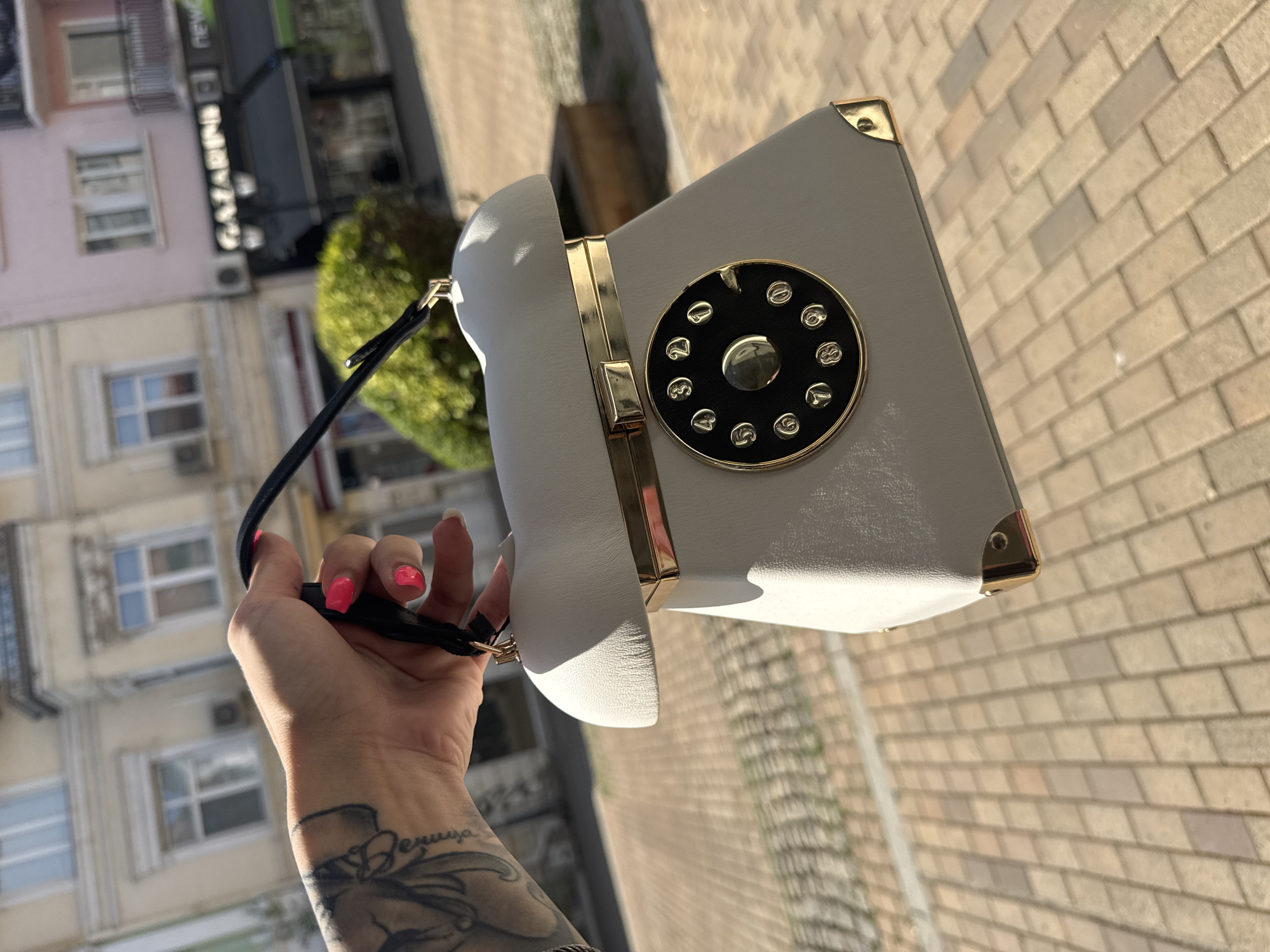 A person's hand, with bright pink fingernails and a tattoo on their arm, holds a novelty handbag designed to resemble a vintage rotary telephone. The handbag is light gray with gold accents, including a decorative dial with numbers and a central gold button, and gold corner protectors. It has a black handle. The handbag is held in front of a blurred background of buildings with an architectural style typical of European cities, possibly Ruse, Bulgaria, given the style of the buildings and the cobblestone pavement. The lighting suggests it is a sunny day.