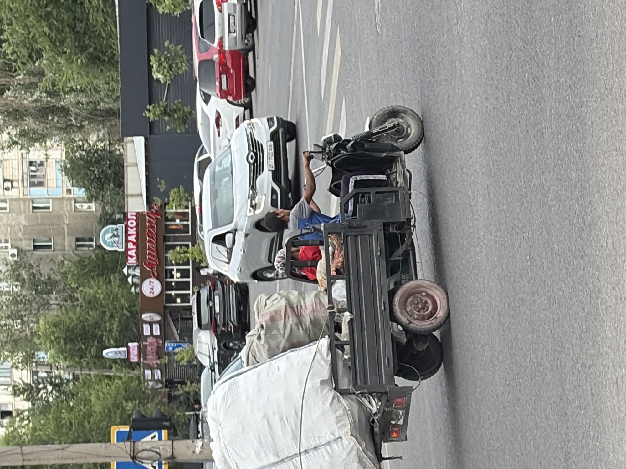 A three-wheeled utility vehicle, loaded with two large white bags and one brown bag, is being driven by a male individual in a city environment. The driver, wearing a gray shirt, is seated and operating the vehicle. Another person, wearing a red shirt, is visible in the passenger area behind the driver. The vehicle is on a paved road, likely an intersection, with other cars present in the background, including a white SUV, a black SUV, a white sedan, and a red car. Buildings and trees form the backdrop of the scene. The overall environment suggests an urban setting.