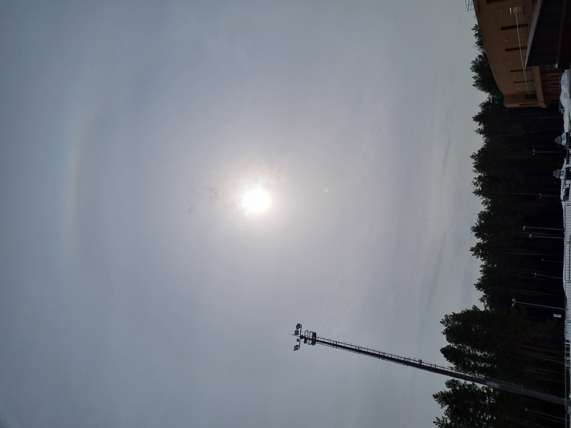 The image depicts a winter outdoor scene in Прикамский, Russia, under an overcast sky featuring a bright, diffuse sun and a faint solar halo. In the left midground, a tall, metal floodlight tower with a ladder extends upwards, crowned with multiple light fixtures. A dense line of dark conifer trees forms the horizon across the midground. To the right, a multi-story building with light brown brickwork, dark vertical windows, and a dark roof is partially visible. The ground is covered in snow. White metal fencing is present in the lower foreground. In the lower right, partially obscured by the fence, at least two snowmobiles are parked on the snow. No individuals or specific actions are discernible.