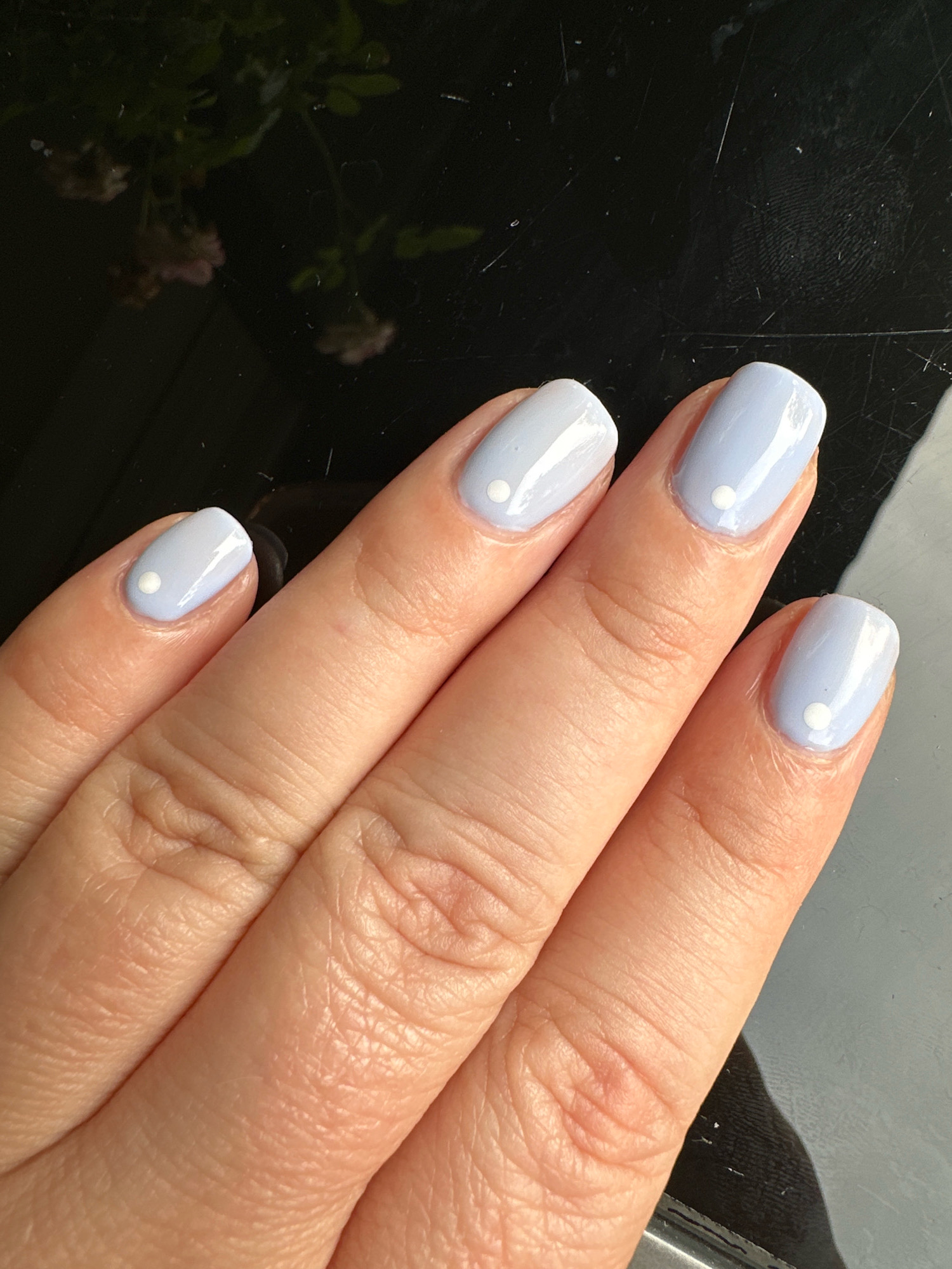 The image shows a close-up of a person's hand with light blue nail polish adorned with small white dots on each fingernail. The hand is positioned over a dark, reflective surface, possibly a table. In the upper left corner of the image, out of focus, is a reflection of some pink flowers and green foliage. The scene does not provide any specific geographical markers to pinpoint a location within Krefeld, Germany.