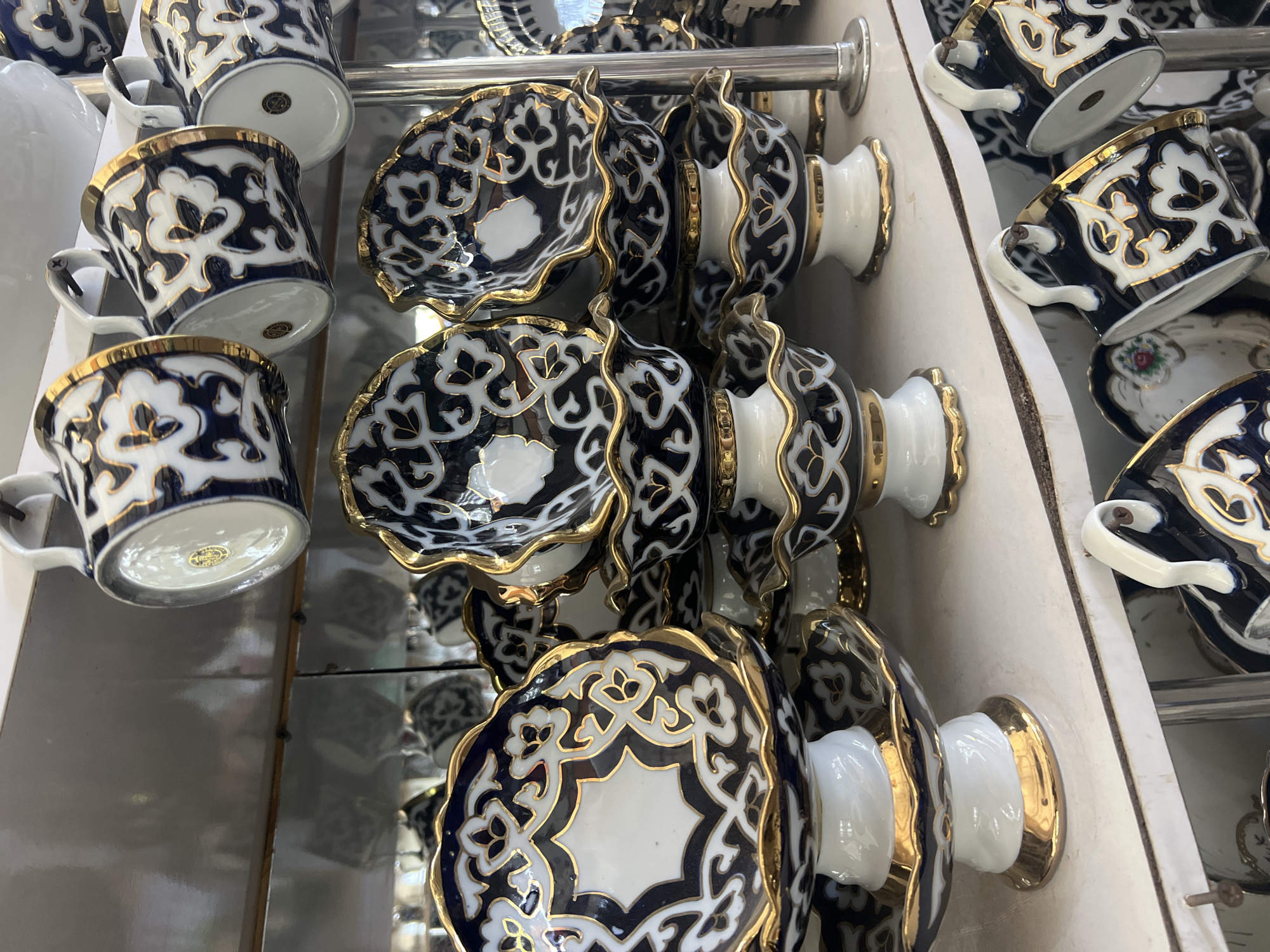 In Eshanguzar, Uzbekistan, on a white shelf, a collection of blue and white ceramic tea cups and saucers with gold trim are displayed. The cups hang from hooks on the shelf above, while the saucers are stacked on the shelf below. The intricate patterns on the ceramics are characteristic of traditional Uzbekistani artistry. The background appears to be a store or market setting with reflective surfaces, suggesting a retail environment.