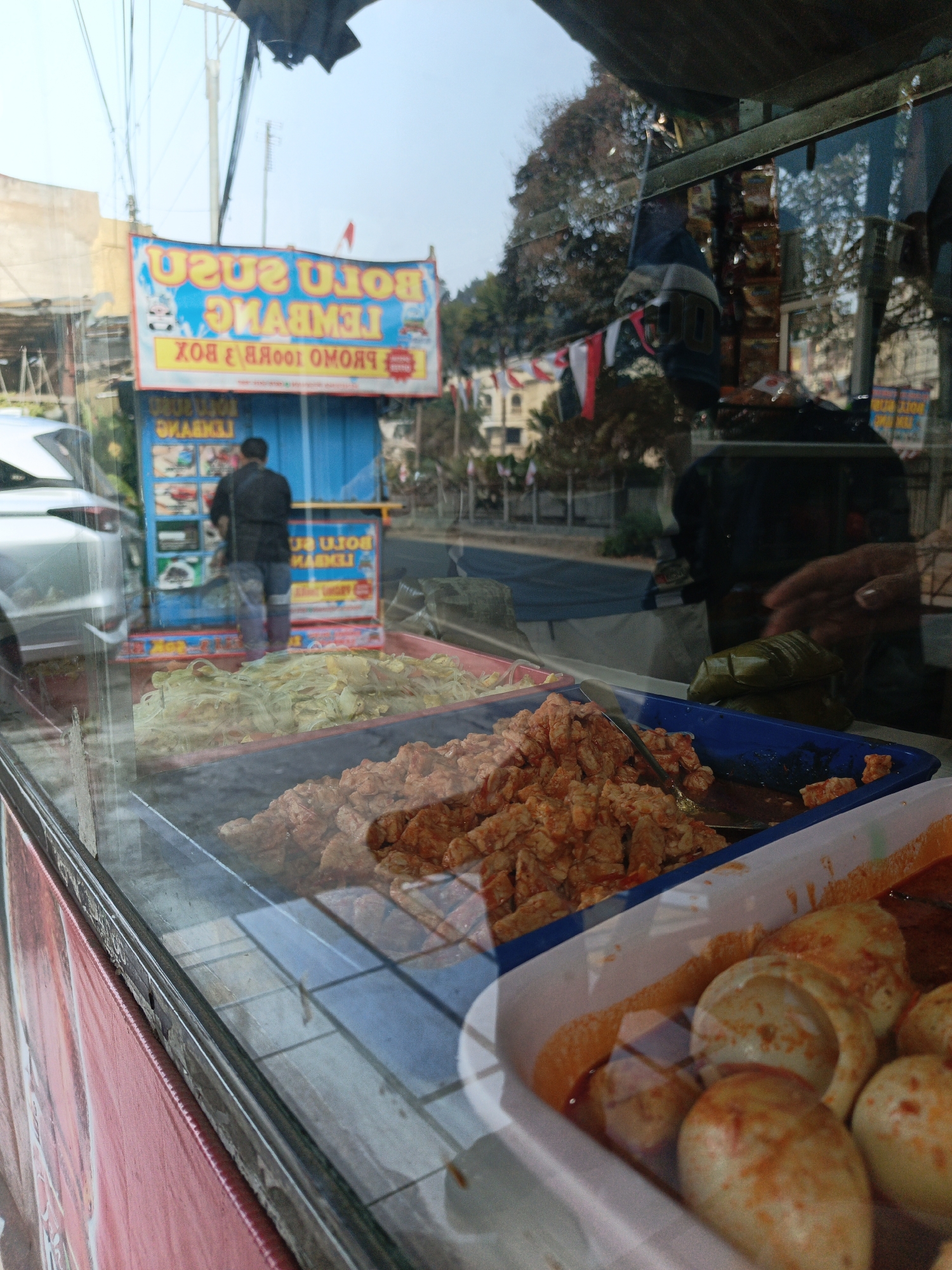 A street food stall is pictured in Cibogo, Indonesia. The stall is selling various food items, including what appears to be noodles, fried tofu, and boiled eggs. A man in a black shirt and blue jeans is visible inside the stall, interacting with the food. Reflections in the glass display show trees, buildings, and flags. The stall has a blue and yellow sign with text in Indonesian. The image is taken from the perspective of a customer looking through the glass display case.