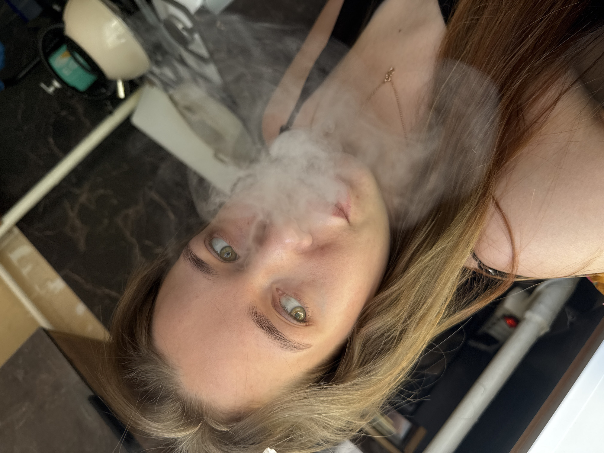A rotated close-up portrait features a young woman exhaling a prominent cloud of white smoke or vapor. Her light brown hair cascades around her face and shoulders, and her light green eyes are fixed directly on the viewer. Her complexion is fair, and a delicate gold chain with a small pendant rests on her chest, visible above a dark, thin-strapped garment.
The cloud of smoke emanates from her mouth, partially obscuring her nose and the central part of her face, giving the image a soft, diffused quality. The composition is dramatically angled, with the woman's head tilted significantly, suggesting an unconventional photographic perspective.
The background elements point to an indoor domestic setting. To the upper left, part of a white, bowl-shaped object with a teal interior is visible, possibly a humidifier or diffuser. The dominant surface in the background is a dark, mottled countertop or table. Faint glimpses of other household items, such as a vertical white pipe-like structure and a subtle red light, can be seen in the periphery. No visible text is present in the image. The scene is intimate, focusing primarily on the woman and the vapor, set within what appears to be a home in Chaikovsky, Russia.