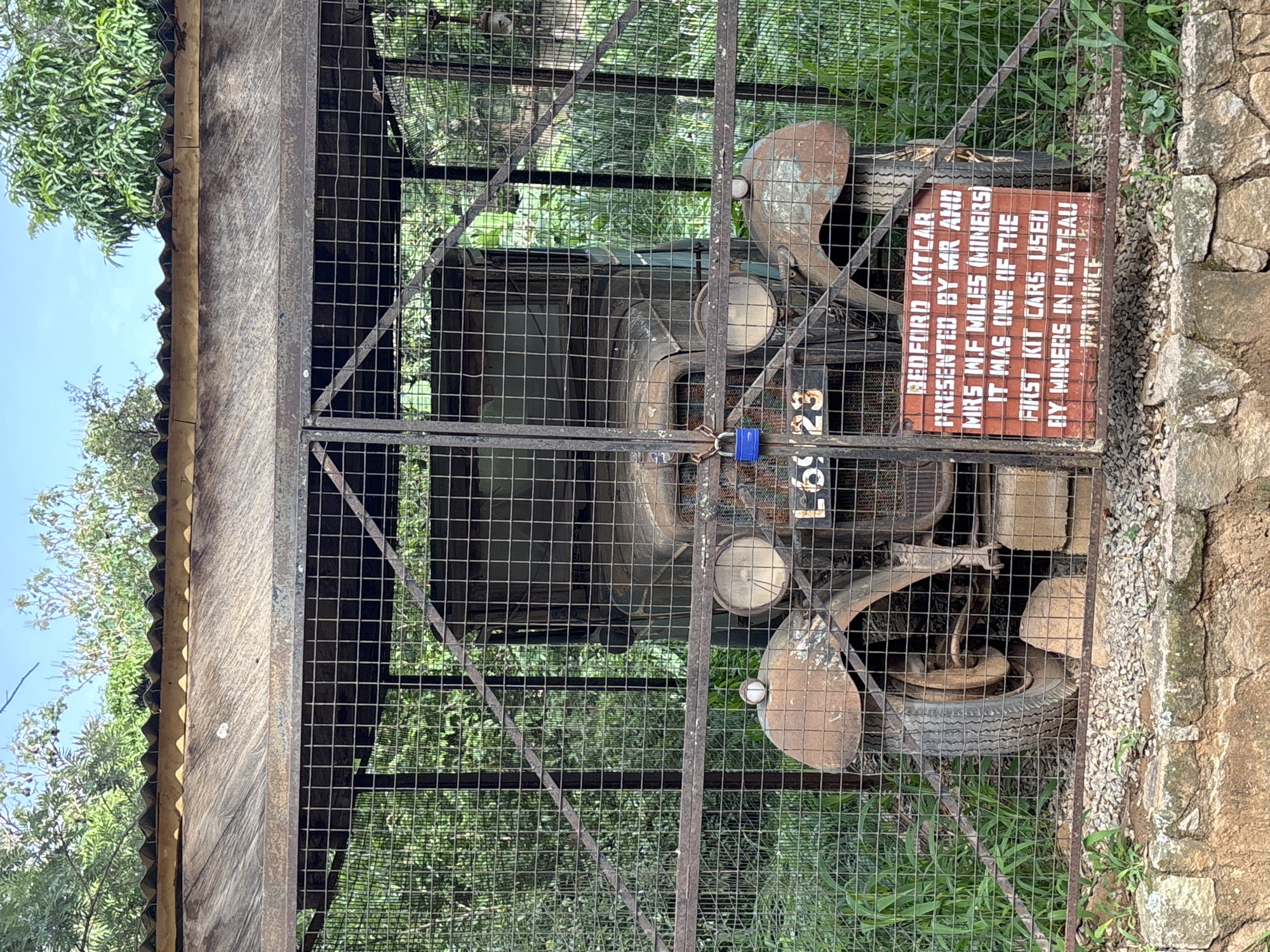 The image shows a rusted, vintage Bedford kit car enclosed in a metal cage with a corrugated metal roof. The car has two large circular headlights and a license plate that reads "L6923". A blue padlock is attached to the cage door. A red sign in front of the car, partially obscured by the cage, reads "BEDFORD KITCAR PRESENTED BY MR AND MRS W.F. MILES (MINERS). IT WAS ONE OF THE FIRST KIT CARS USED BY MINERS IN PLATEAU PROVINCE". The car is situated outdoors, surrounded by dense green foliage and trees, under a bright sky. The ground in front of the cage is covered with small stones and larger, rough-hewn stones. This scene is located in Jos, Nigeria, likely at a historical or museum site dedicated to the mining heritage of the Plateau Province.