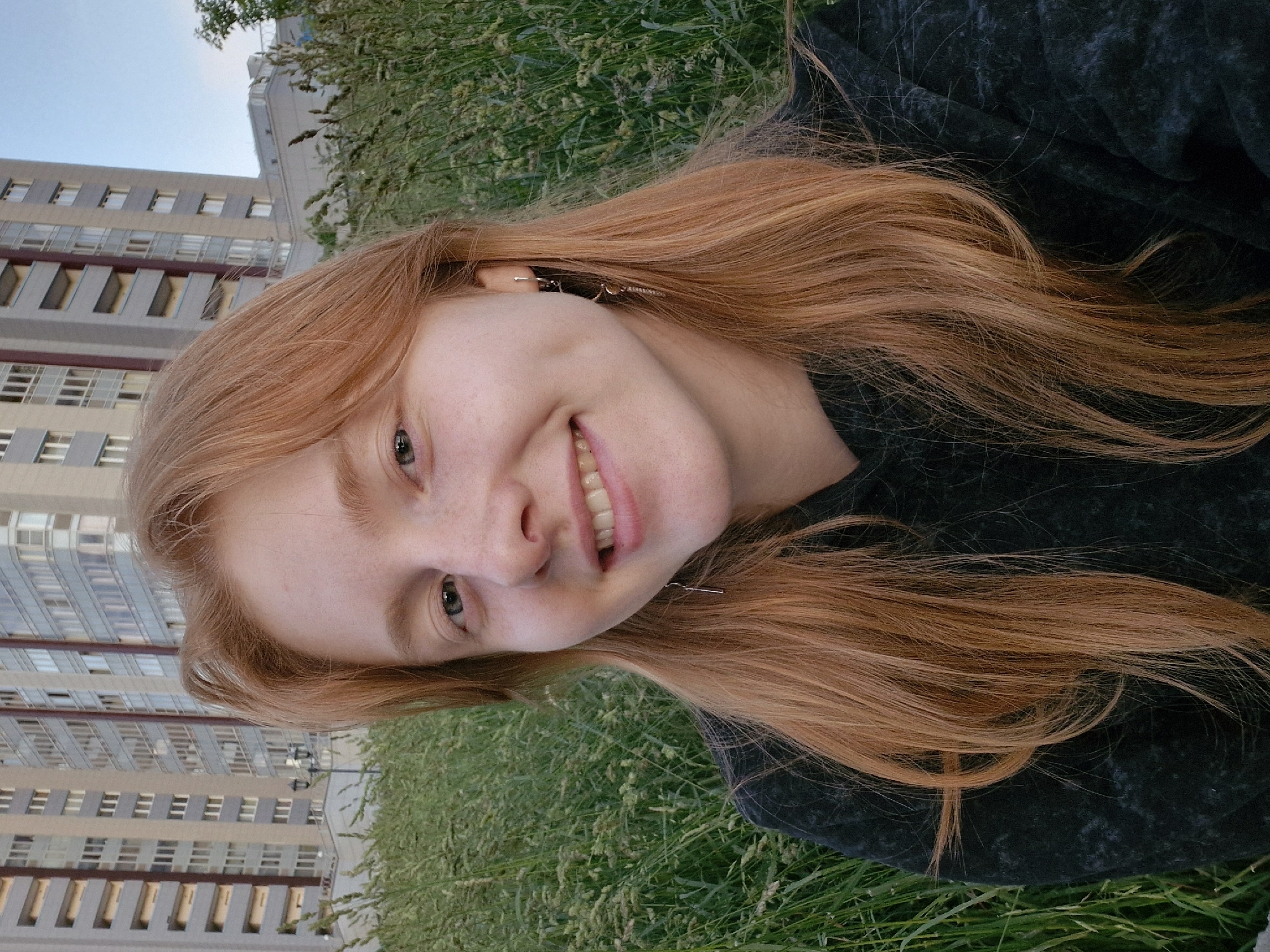 A young woman with long, auburn hair smiles into the camera. Her hair is spread across a dark surface, possibly a blanket, with green grass and some dried vegetation visible in the foreground and background. Behind her, a modern, multi-story building with many windows is visible, suggesting an urban or suburban setting. The sky appears clear and blue, indicating it's daytime and likely good weather. The woman is wearing a dark top. Her expression is happy and relaxed, with her eyes open and a slight grin. She has a few freckles visible on her cheeks. Her hair is a prominent feature, cascading around her. The image is shot from a low angle, with the woman's head tilted to her right.