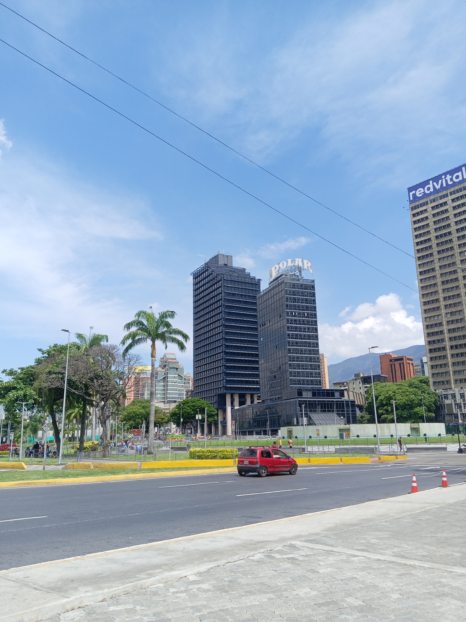 The image captures a street scene in Caracas, Venezuela. Prominent in the frame are two identical, tall, dark-colored buildings labeled "POLAR" on top. Another building, labeled "redvital" is visible to the right. A red vehicle drives on the road in front of the buildings, and pedestrians are visible further away. A yellow curb separates the road from a green space with trees. The sky is blue with some clouds, and power lines stretch across the top of the image. The pavement in the foreground is gray. Traffic cones are positioned on the right side of the road.