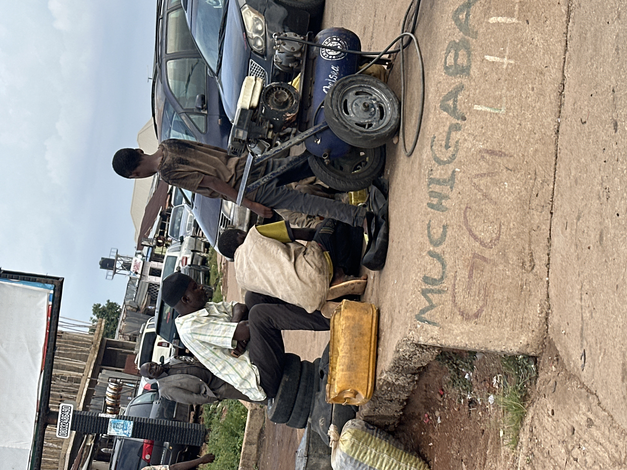 In Gura Top, Nigeria, three individuals are engaged in activities around what appears to be a roadside repair or service area. The scene is set outdoors, with a paved surface and a backdrop of parked vehicles and makeshift structures.
The primary subjects are three men. One man, wearing a brown, tie-dyed t-shirt, is positioned in the foreground, leaning over an air compressor. Another man, dressed in a light-colored, checkered shirt and a black cap, sits on a stack of tires. The third man, wearing a light brown or tan garment, is seated next to the man on tires.
Prominent objects include a blue air compressor with a hose, a stack of old tires serving as seating, and a yellow plastic jerrycan. A car is partially visible in the background. The ground is a rough, paved surface with some dirt and vegetation.
The activity seems to be focused on car maintenance or tire inflation, suggested by the presence of the air compressor and tires. The overall atmosphere appears to be one of daily labor in a bustling, informal setting.
Notable details include the overcast sky, suggesting a cloudy day. The graffiti on the ground, though partially obscured, provides a clue to the location.
Visible text includes "Chelsea" on the air compressor. On a billboard-like structure to the left, there is a phone number, "08032485450," and what looks like "GURA TOP." There is also text written in chalk or paint on the ground which appears to spell out "MUCHAGABA" and "CALABAR".