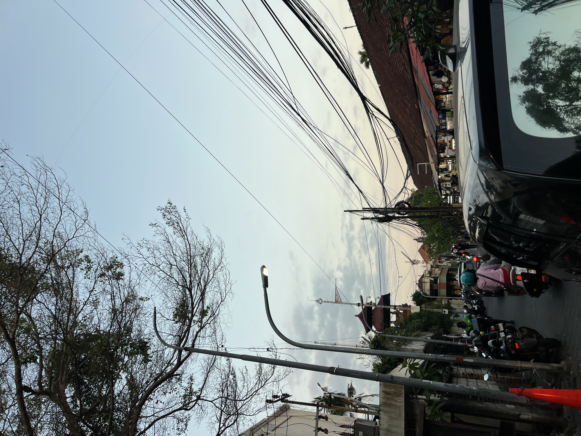 This image depicts a street scene in Kuta, Indonesia, during twilight. The sky is a soft blue with hints of pink and orange from the setting sun, partially obscured by scattered clouds and a dense network of overhead electrical wires. The foreground is dominated by the rear of a dark-colored SUV, its roof antenna visible. To the left, a street lamp with its light on illuminates a portion of the sidewalk and the side of a building. A traffic cone sits on the sidewalk near the building.
Further down the street, a line of motorcycles is parked, and several people are on them or walking. One individual on a motorcycle is wearing a pink jacket and a teal helmet. Other buildings line the right side of the street, featuring tiled roofs and what appear to be market stalls or shops with colorful awnings. The architecture suggests a tropical or Southeast Asian setting. The overall impression is a busy urban street transitioning into evening.