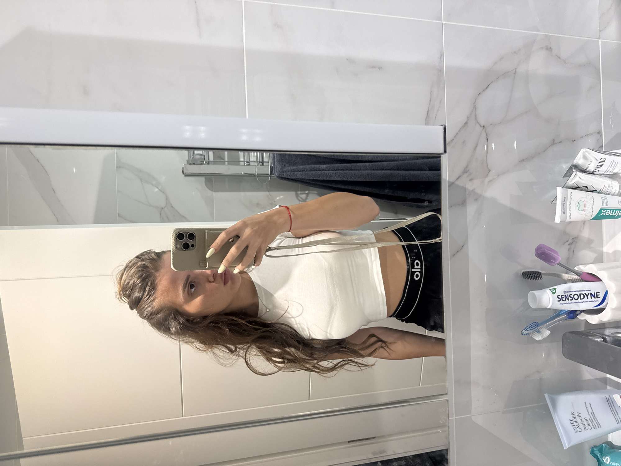 A young woman is taking a mirror selfie in a modern bathroom. She is wearing a white crop top and black shorts with a white logo band. Her long, wavy brown hair is pulled up loosely. In the foreground, a bathroom sink is visible with various toiletries, including toothbrushes, toothpaste, and a face mask tube. The bathroom features white marble tiles and modern fixtures. The scene does not provide any specific information to indicate a location in Throop, United Kingdom.