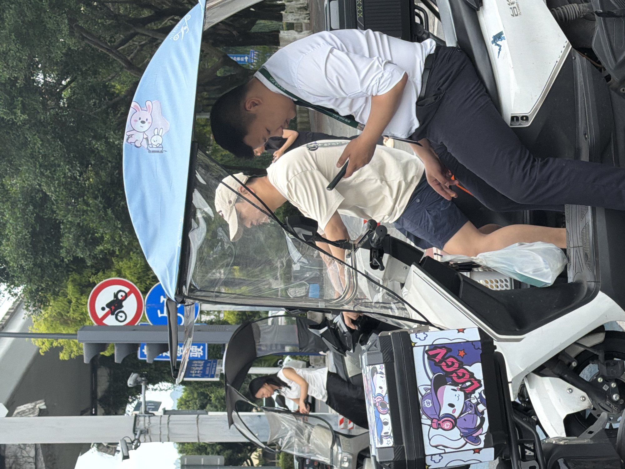 Two men in white shirts are on an electric vehicle. One is operating the vehicle, while the other appears to be making adjustments with a tool. The electric vehicle has a windshield with a cartoon of bunnies on it. The scene is located in Chongqing High-tech Zone, China, as indicated by the street sign visible in the background. The background includes signs indicating "no motorcycles," tree foliage, and overhead structures. Another woman is also driving another vehicle in the same line of vehicles. The battery box of the electric vehicle has a cartoon design on it.