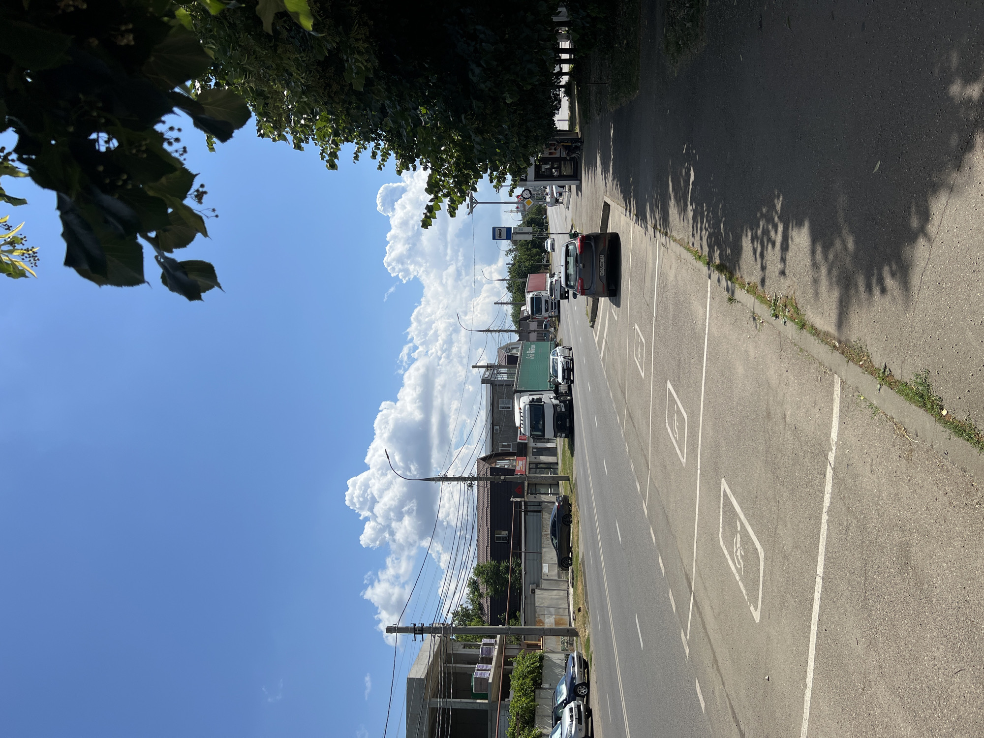 The image shows a street scene in Krasnodar, Russia. The street is paved, with multiple lanes for traffic. There are designated parking spots marked on the side of the road, some of which are marked with symbols indicating disabled parking. Several vehicles are visible, including cars and a large truck. Buildings line one side of the street, with some appearing to be under construction. Trees and foliage are present, casting shadows on the sidewalk and road. The sky is blue with scattered white clouds. A bus stop shelter is also visible on the sidewalk.