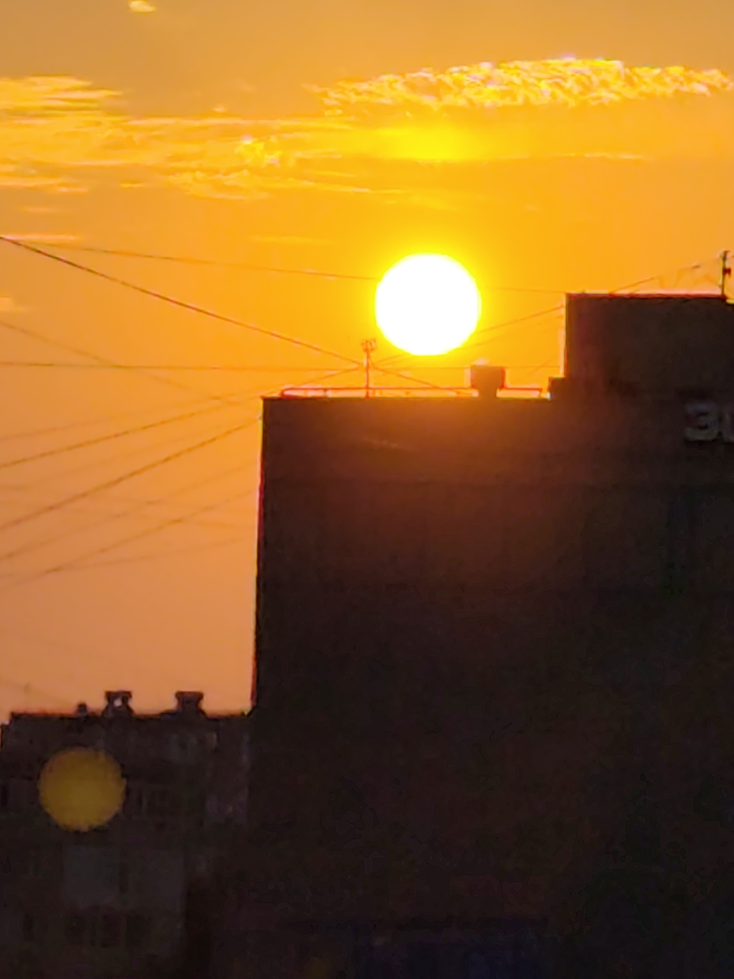 The image depicts a sunset in the city of Aktau, Kazakhstan, casting a vibrant orange and yellow glow across the sky. Silhouetted against the dramatic sky are several apartment buildings, their dark forms contrasting sharply with the bright light. Power lines crisscross the frame, adding a utilitarian element to the otherwise picturesque scene. The sun, a bright, perfect circle, is the focal point, positioned slightly to the right of the center, illuminating the clouds and the upper portions of the buildings. The lower parts of the buildings are obscured by shadow, suggesting the sun is low on the horizon. The overall impression is one of a city bathed in the warm, ephemeral light of a setting sun.