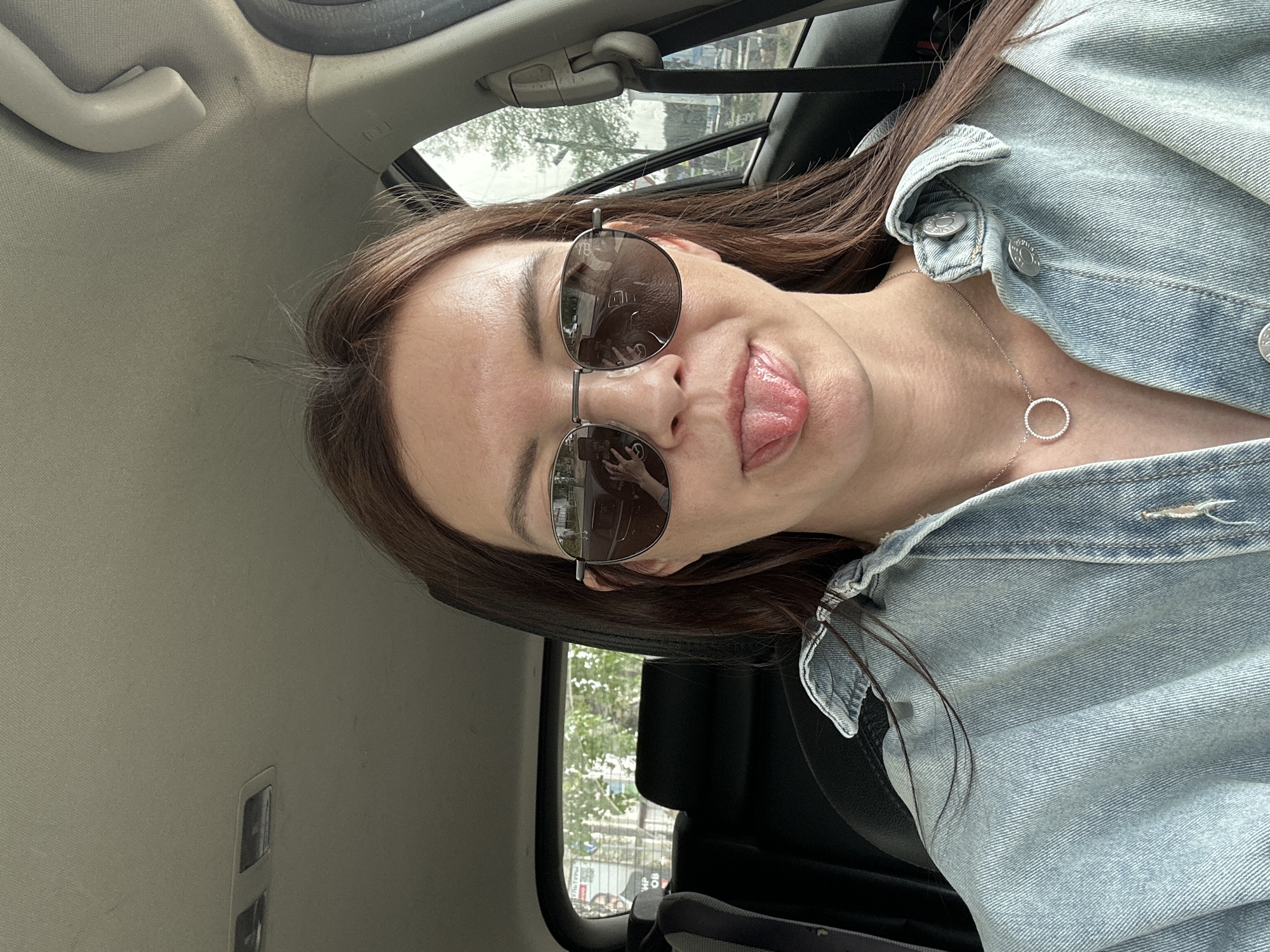 A young woman is in the passenger seat of a car. She is wearing a denim jacket, a necklace, and sunglasses and sticking her tongue out. The car appears to be in Chaikovsky, Russia, as that location is reflected in the sunglasses. Outside the car window, there are trees, buildings, and a street.