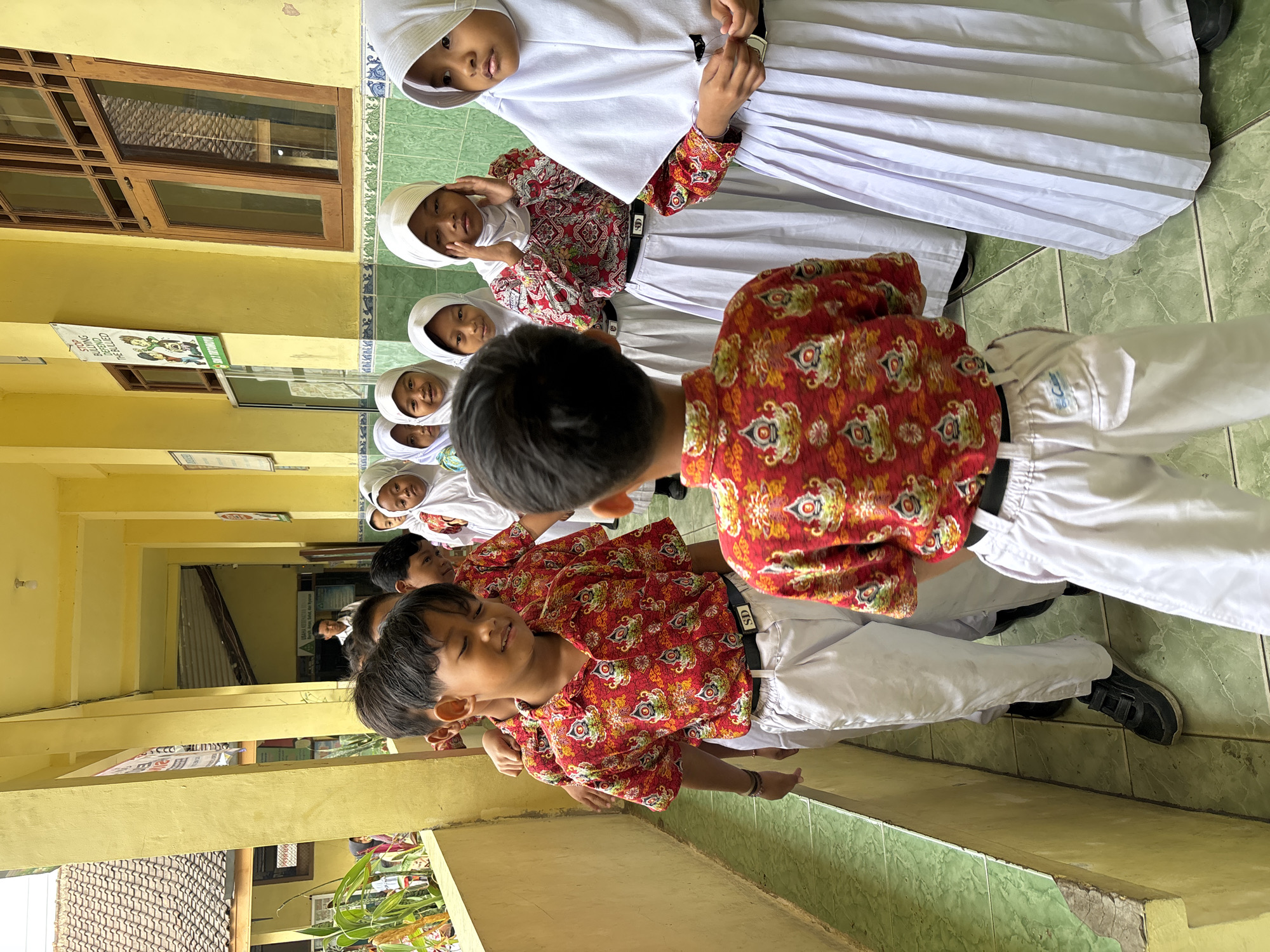 In Tirtomoyo, Indonesia, a group of school children, both boys and girls, are standing in a hallway. The boys are wearing red shirts with a patterned design and white pants. The girls are wearing white headscarves and white skirts, some with patterned tops. The hallway has yellow walls and a green tiled floor. A banner with text and images is visible on the yellow wall. Sunlight is filtering into the hallway from an opening to the left, illuminating some of the children and the environment. The children appear to be in a line, possibly waiting or moving to a different location.