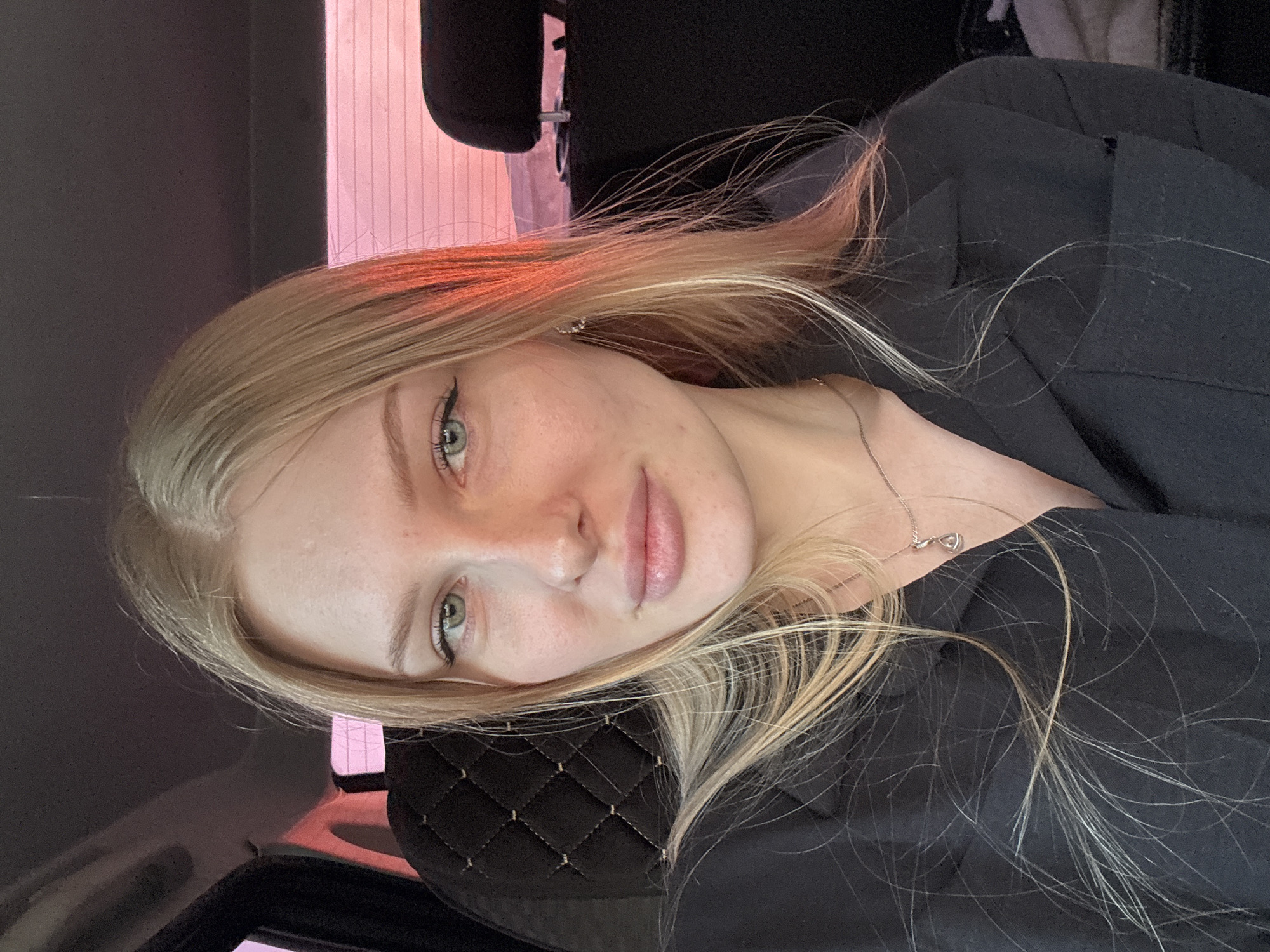 A young woman with long blonde hair is photographed inside a car. She is wearing a black top and a delicate silver necklace. The interior of the car features a black headrest with a diamond-patterned stitching. The lighting in the car is a mix of natural and artificial, with a pinkish hue visible near the window. It is not possible to determine if this image was taken in Кондратово, Russia.