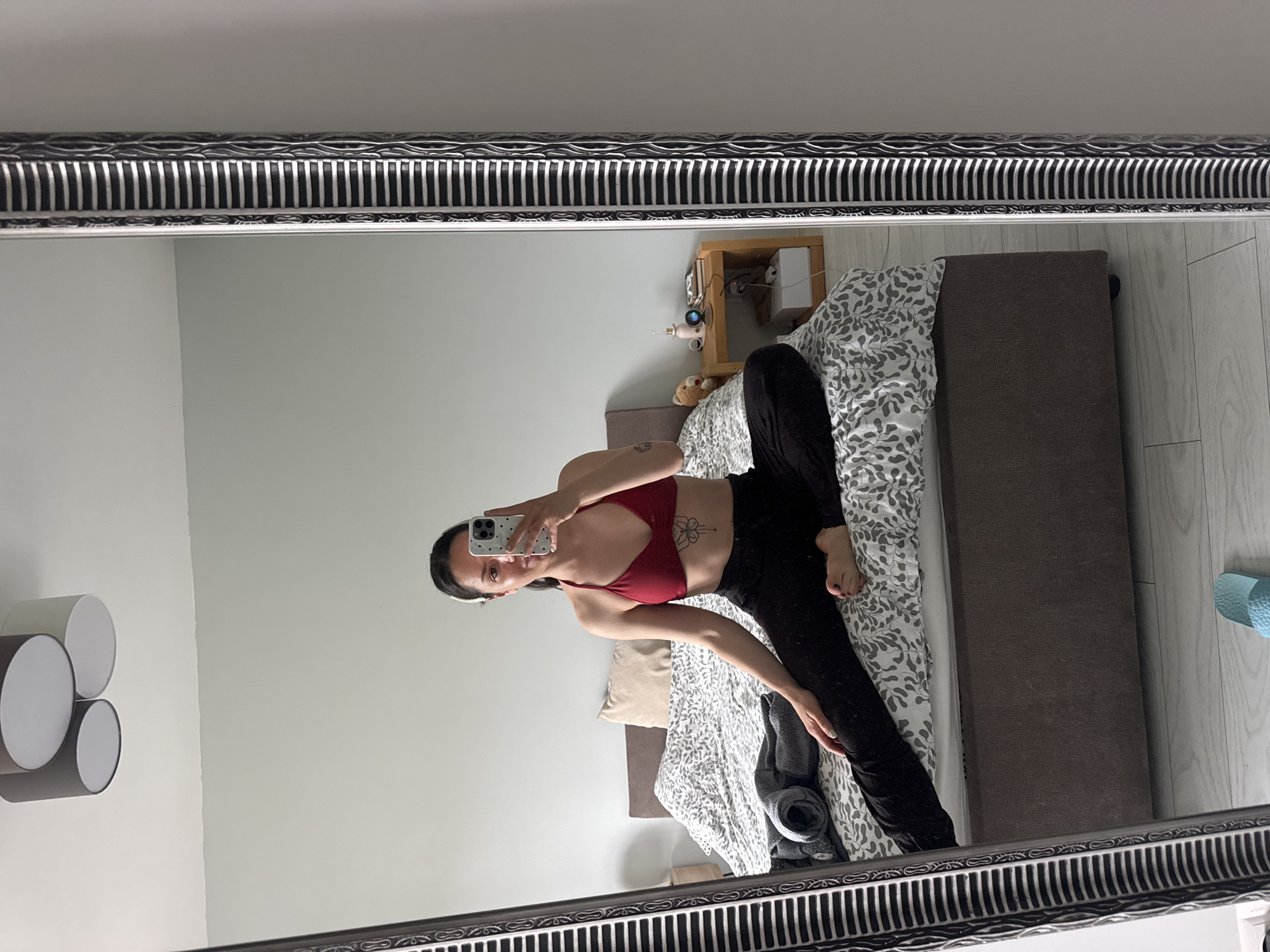 This mirror selfie, taken in a bedroom in Burgas, Bulgaria, captures a young woman sitting on the edge of a bed. She has dark hair, styled with a white headband, and is wearing a red sports bra and black leggings. A floral tattoo is visible on her left side, below her rib cage. She holds a white smartphone with a patterned "Casetify" case, looking directly at her reflection in the mirror to take the photo.
The bedroom features light-colored walls and a light wooden floor. The bed is covered with a white duvet adorned with a grey leaf-like pattern, and a beige pillow is partially visible. To the right of the bed, a wooden nightstand holds a small white electronic device, possibly a baby monitor or security camera, along with a small teddy bear and some cables. On the left wall, a modern light fixture with four circular elements is mounted. A blue mat or towel is visible on the floor near the bed, and a rolled-up dark blanket or garment lies on the bed. The mirror itself is encased in an ornate silver frame featuring intricate black and silver decorative patterns. The scene is brightly lit, suggesting it is daytime.
